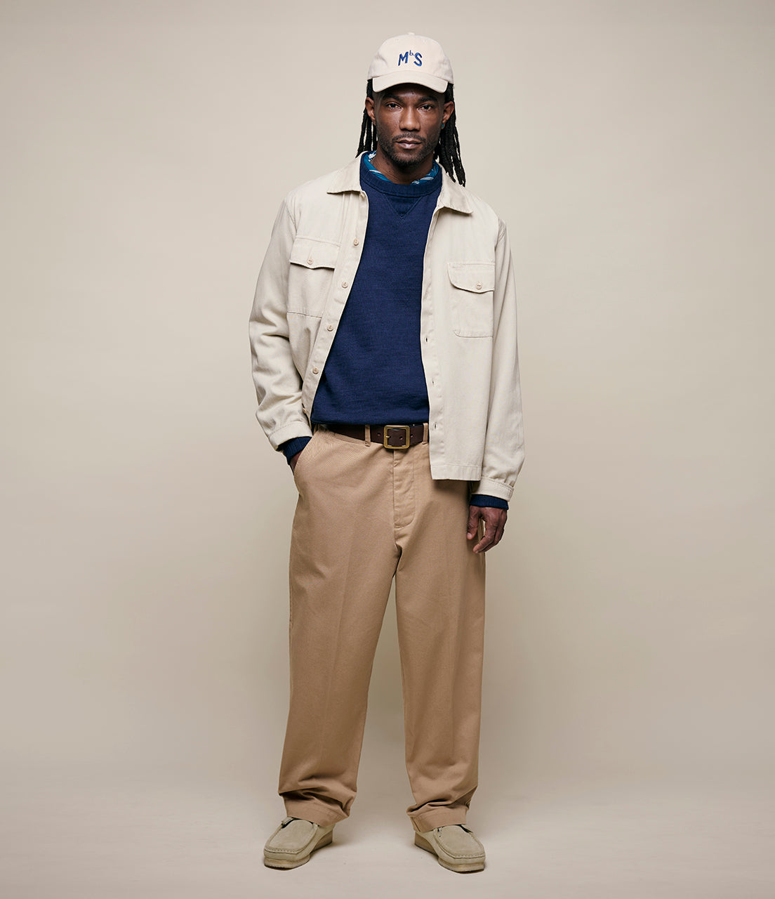 Man wearing a white jacket, blue sweater, beige pants, and a cap on a plain background