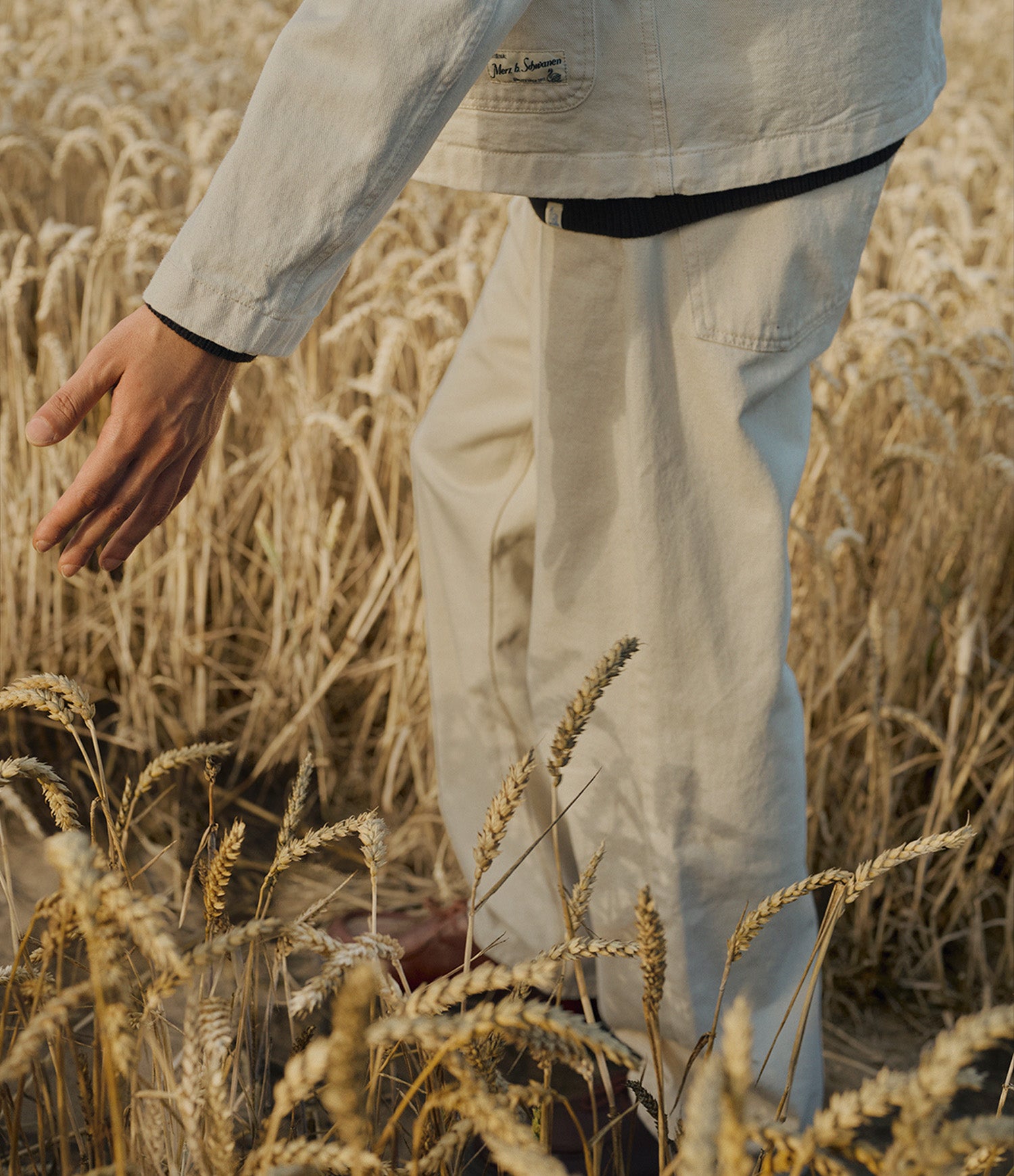 A person standing in a golden wheat field wearing light beige twill pants and a matching light beige jacket, with one hand reaching toward the wheat stalks.