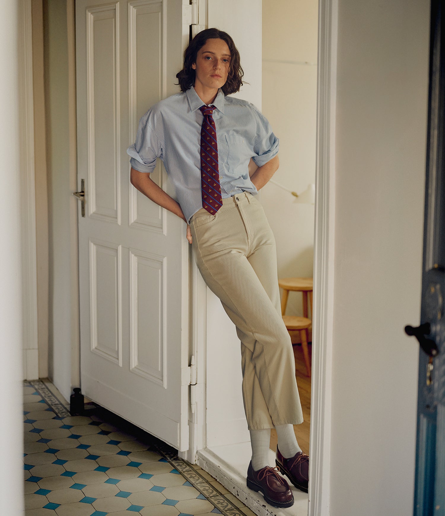 A person leans against an open white door in a hallway, wearing a light blue button-up shirt, a red patterned tie, beige trousers, white socks, and dark brown loafers.