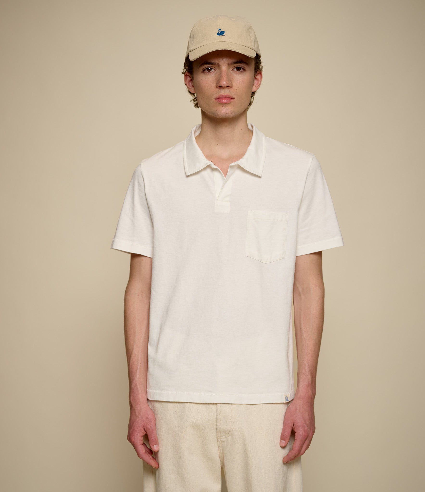 Person wearing a beige cap and white polo shirt against a beige background