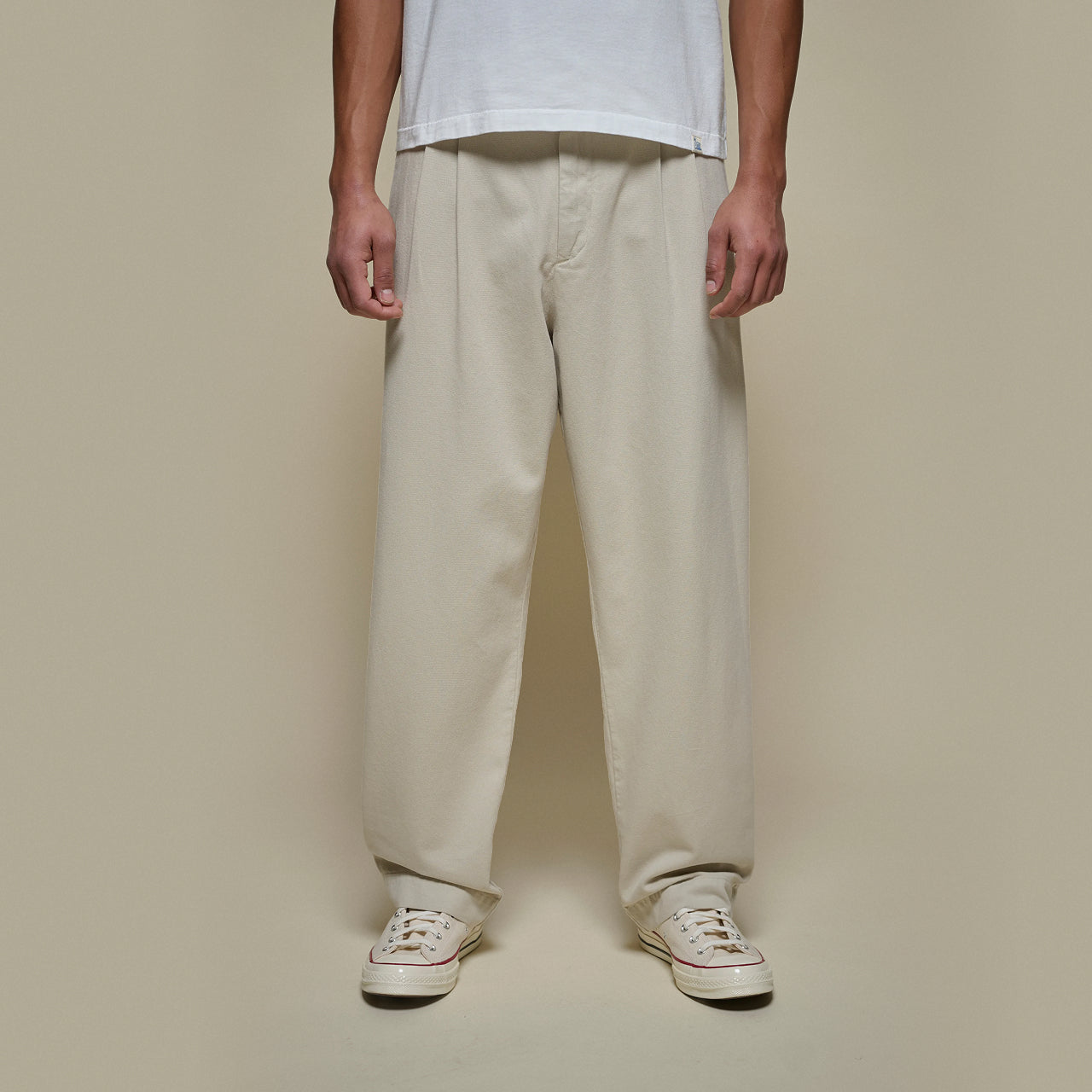 Person wearing beige  pleated pants and white sneakers against a plain background