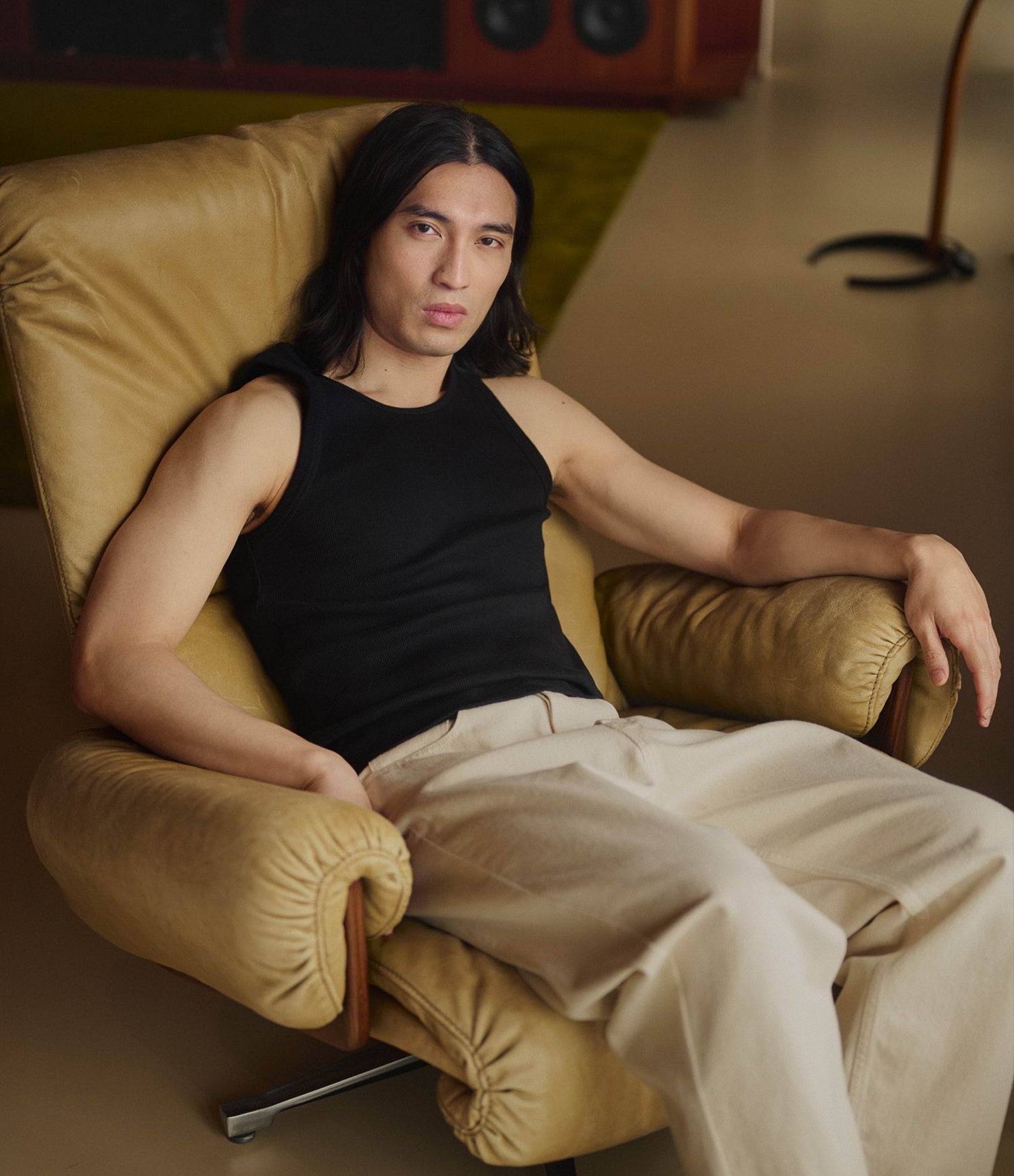 A man with long dark hair sits reclining in a tan leather armchair, wearing a black sleeveless tank top and light beige pants in a softly lit indoor setting.