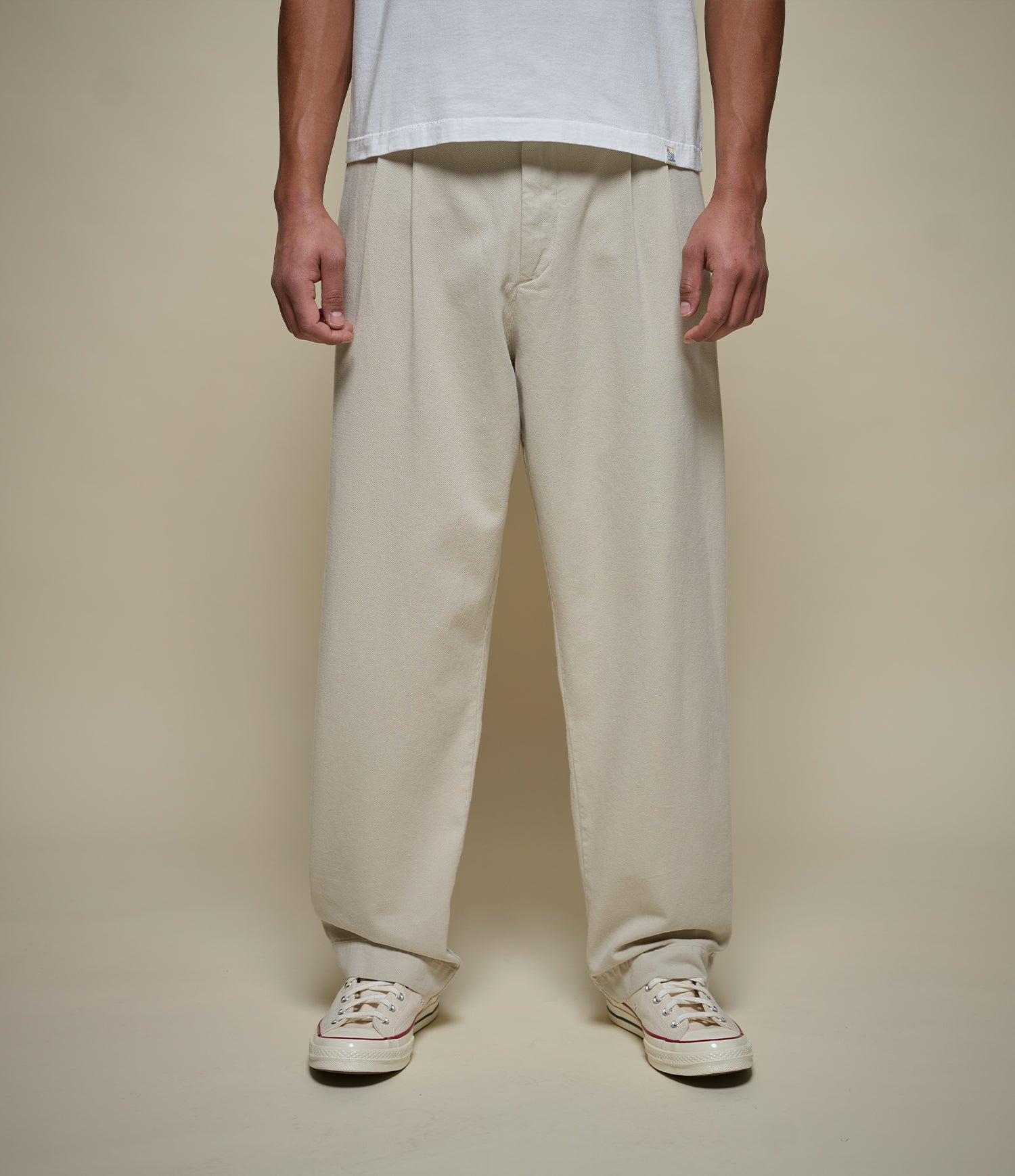 Person wearing beige pleated pants and white sneakers on a plain background