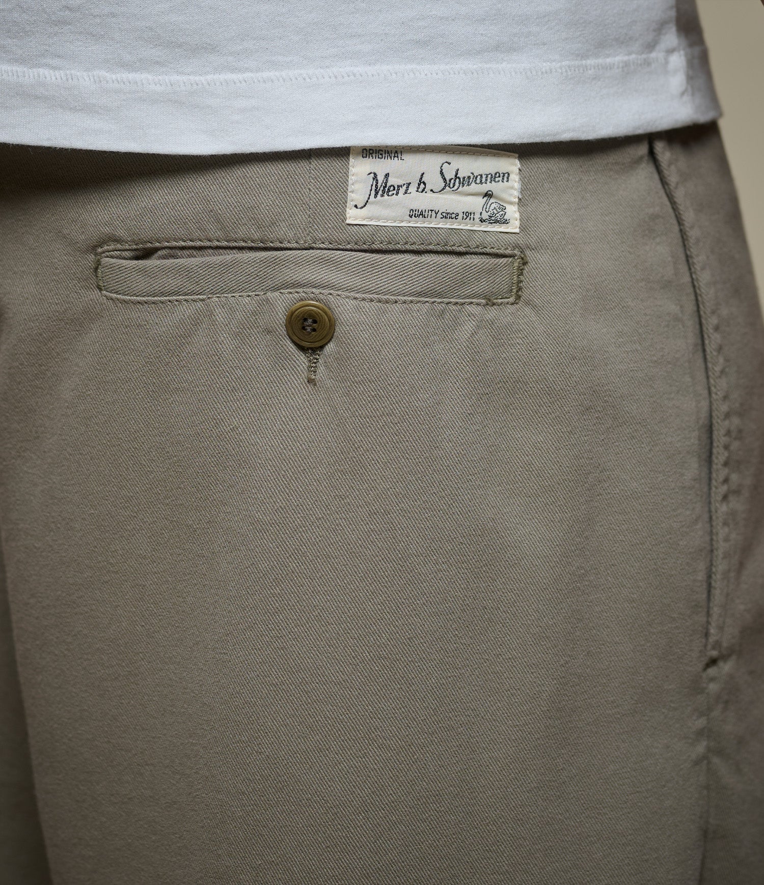 Close-up of a green pants pocket with a button and a brand Merz b. Schwanen label on a beige background