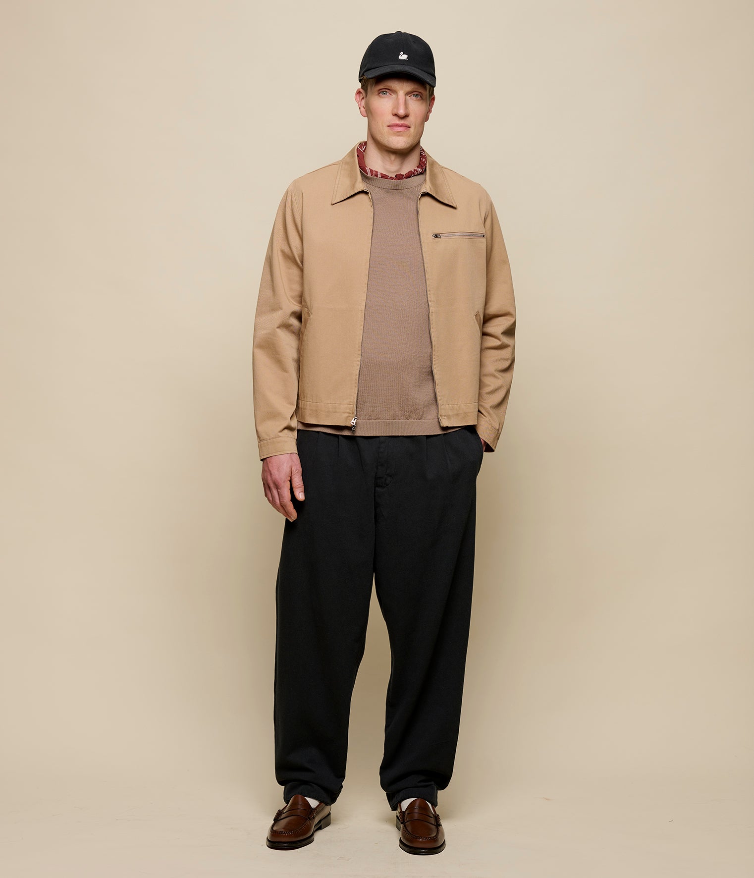 Man wearing a beige jacket, black pants, and a black cap against a beige background