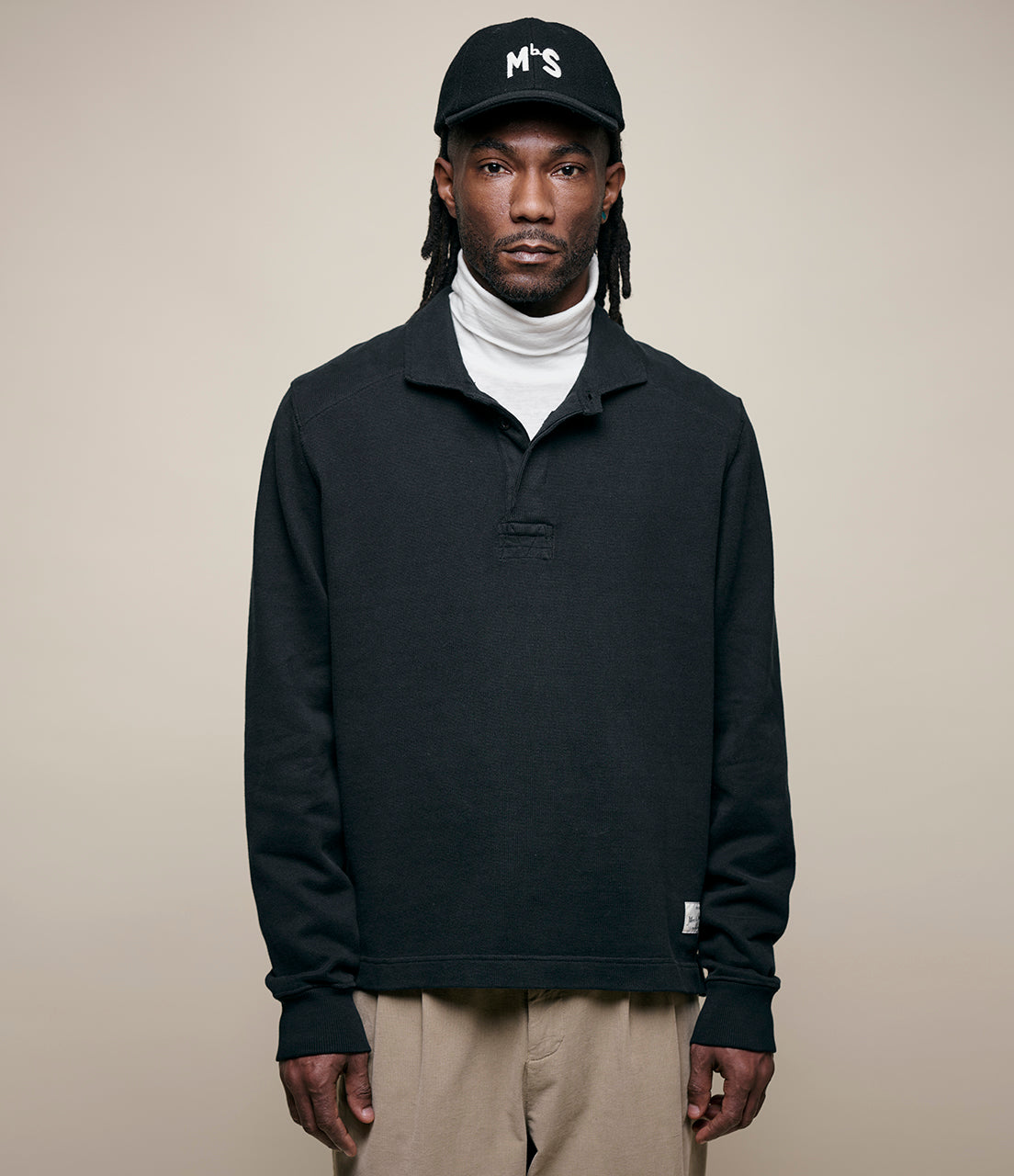 Man wearing a black rugby shirt, white turtleneck, beige pants, and a black cap with 'MbS' logo on a beige background