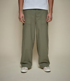 A person wearing loose, wide-leg olive green cargo pants with front pockets, paired with a white t-shirt and white sneakers, standing against a plain beige studio background.