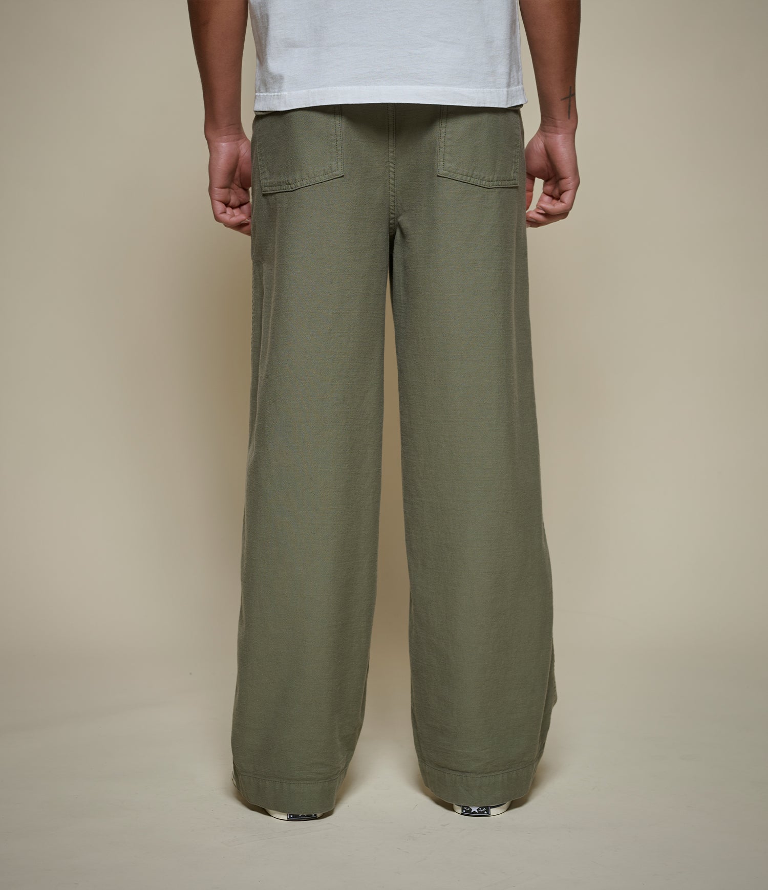 A person shown from the back wearing relexed-fit olive green pants and a white t-shirt, standing against a plain beige studio background.