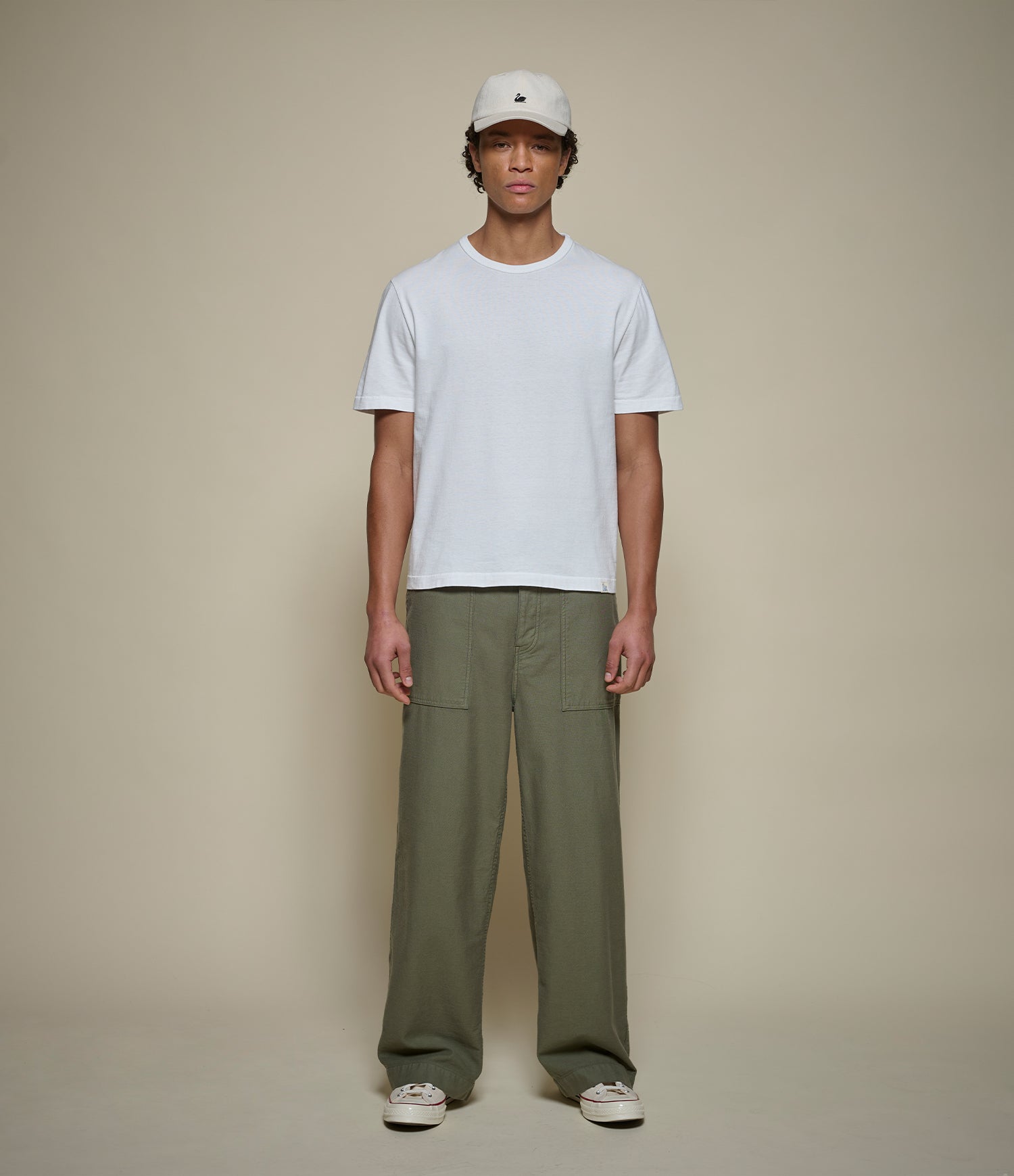 A model stands against a plain beige studio background wearing a white short-sleeve crewneck t-shirt, olive green wide-leg fatigue pants, a white baseball cap, and white sneakers.