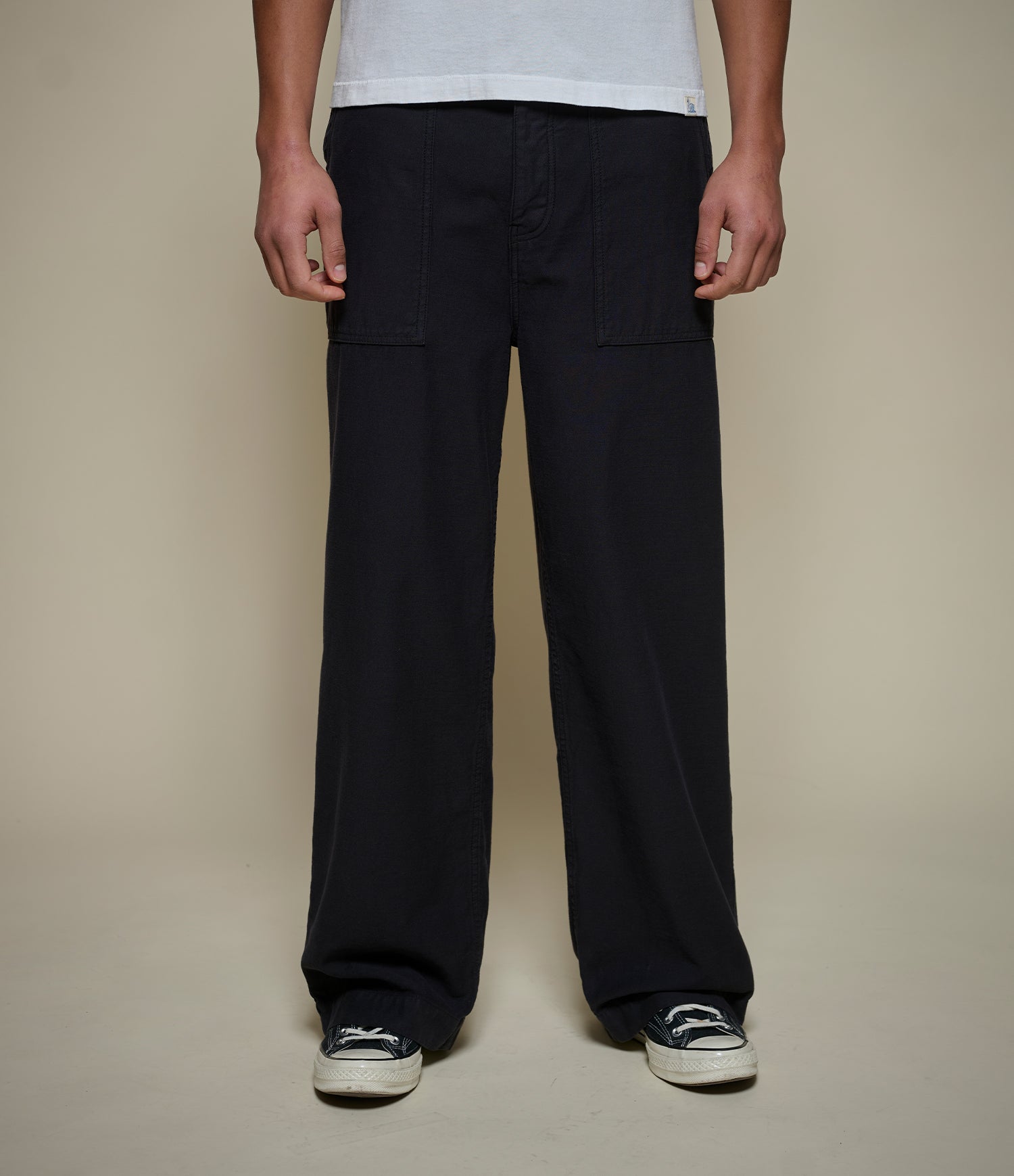 A person wearing black wide-leg cargo pants with front flap pockets, paired with a white t-shirt and black-and-white sneakers, shown from the waist down against a plain beige studio background.