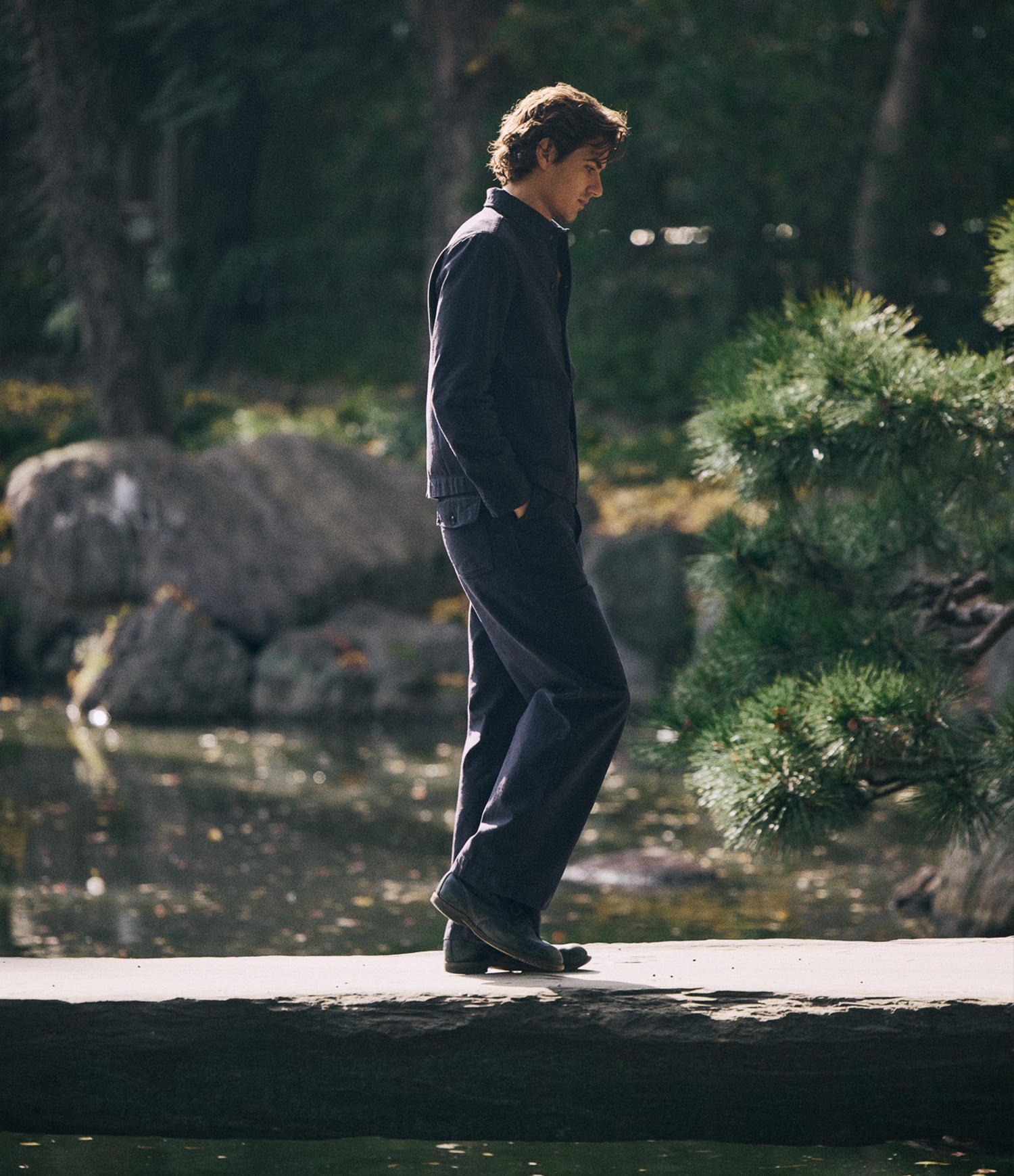 A man in a black sateen canvas jacket and pants walks along a stone path in a serene Japanese garden with rocks, water, and greenery.