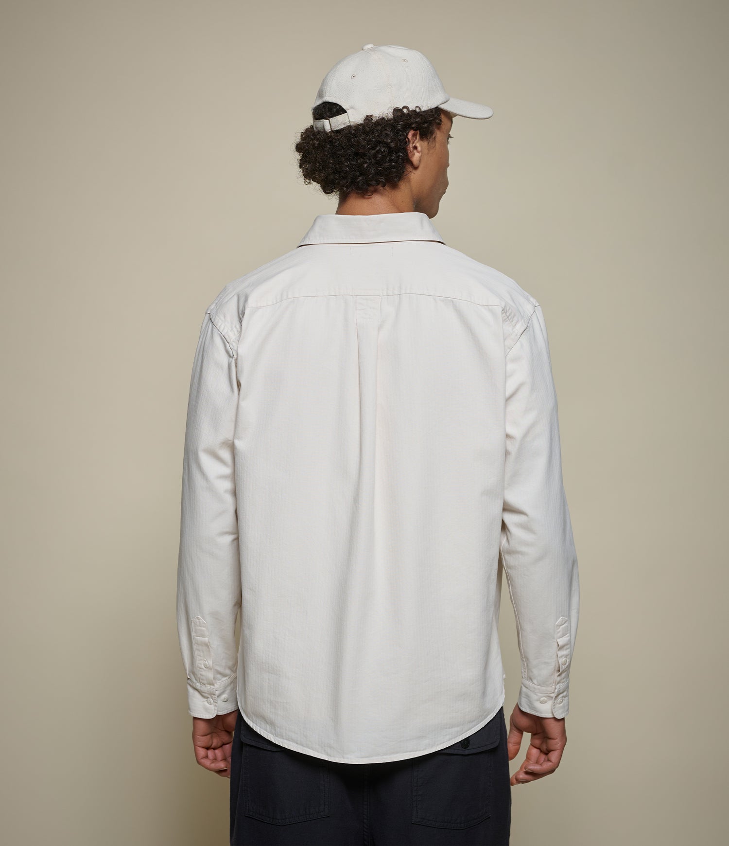 A man viewed from behind wearing a white long-sleeve button-up shirt and a white baseball cap, standing against a plain beige studio background.