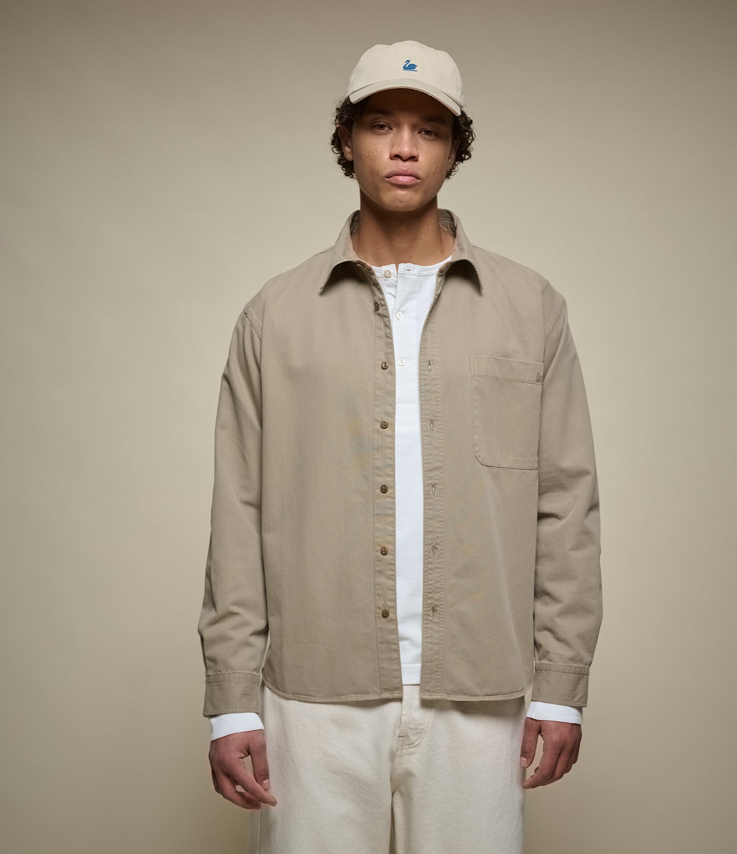 A man wearing a beige button-up herringbone overshirt over a white t-shirt, paired with cream pants and a beige baseball cap, standing against a solid beige background.
