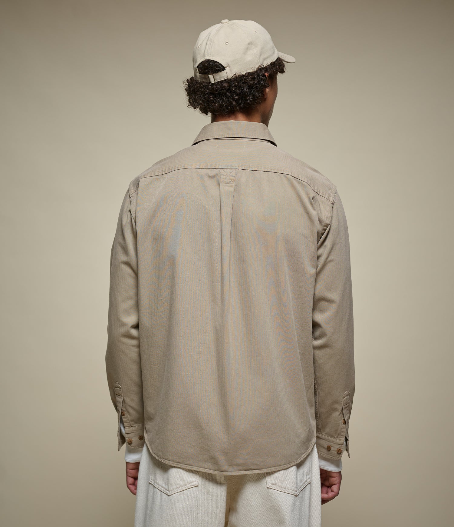 A man viewed from behind wearing a light khaki long-sleeve button-up shirt, white pants, and a beige baseball cap against a solid beige studio background.
