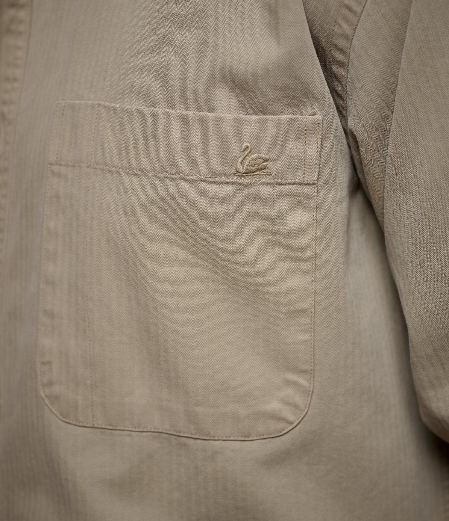 Close-up of a khaki herringbone textured shirt pocket with a small embroidered swan logo on the upper left corner.