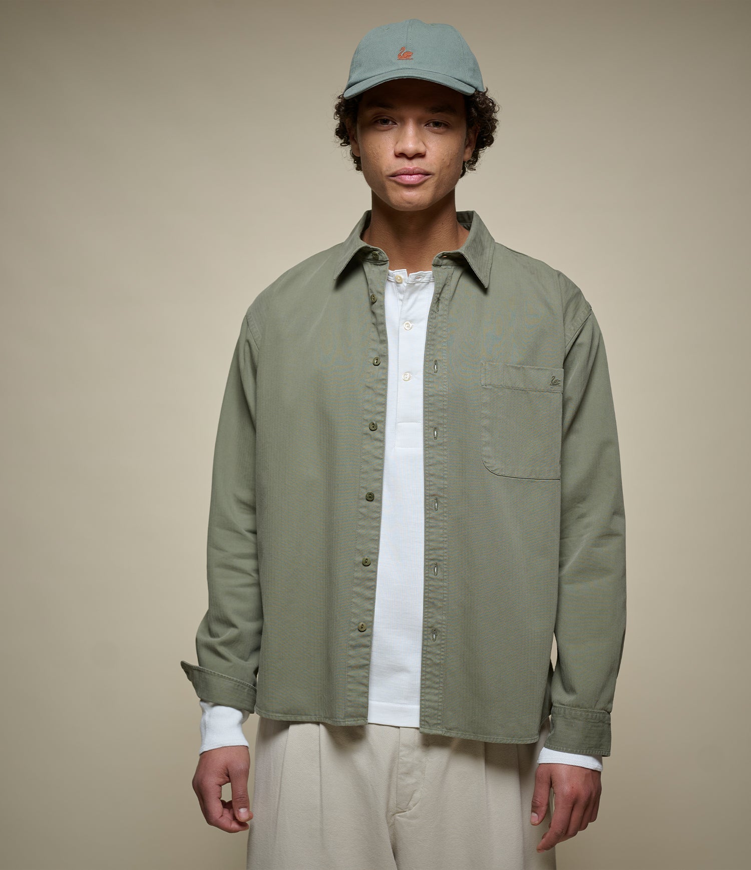 A man wearing a sage green button-up overshirt over a white t-shirt, paired with beige trousers and a matching sage green baseball cap, set against a plain beige studio background.