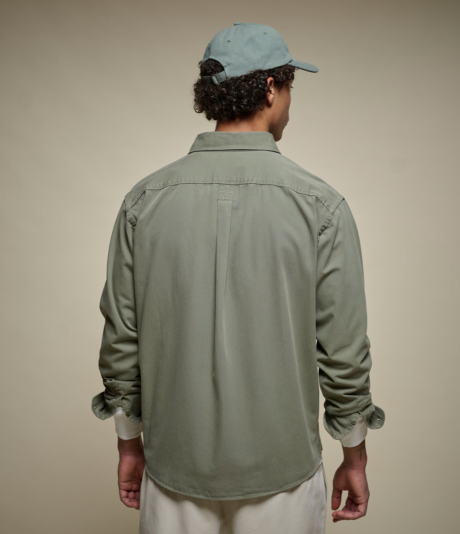 A man viewed from behind wearing a sage green long-sleeve button-up shirt, cream pants, and a light blue baseball cap against a solid beige background.