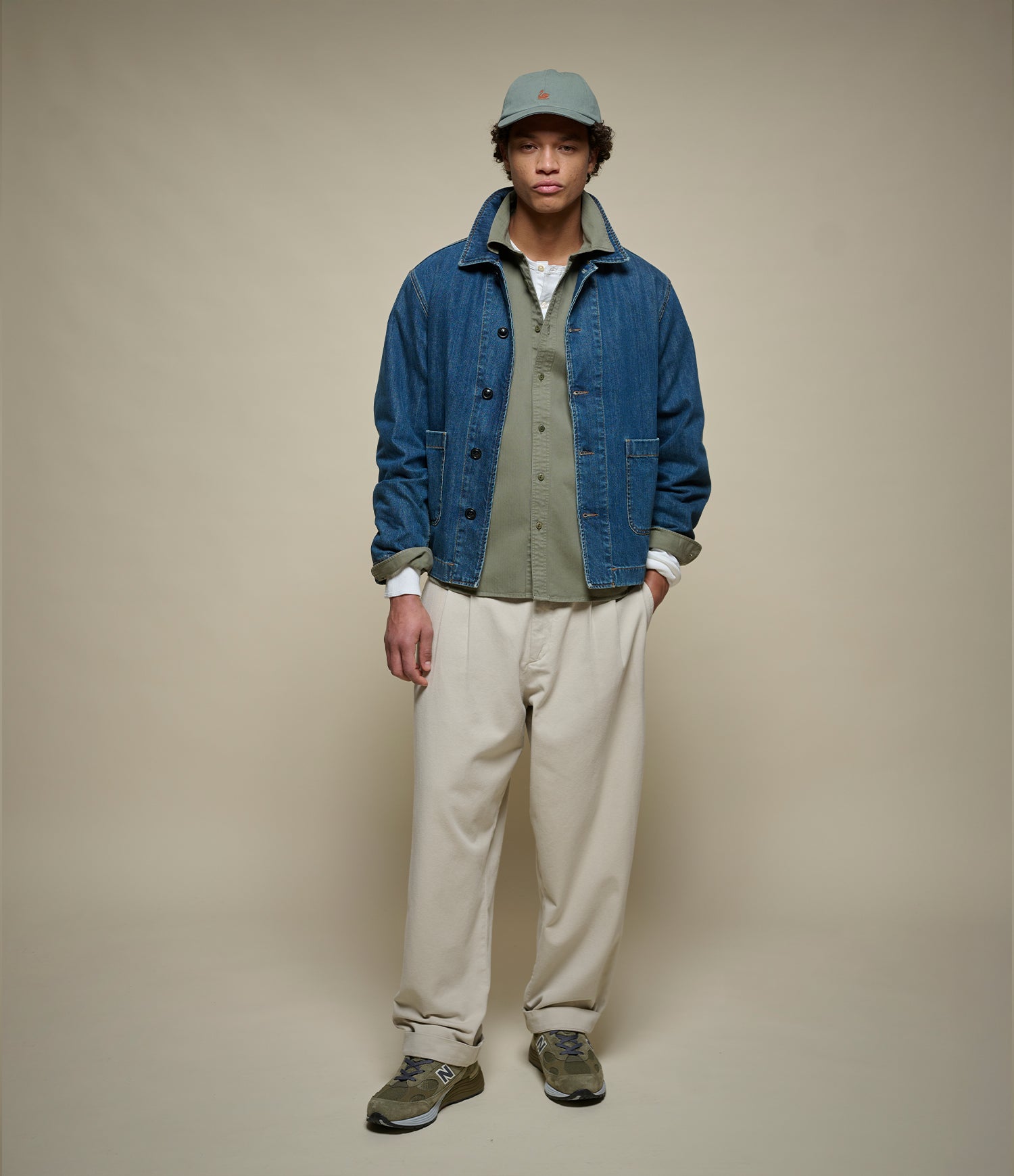 A model stands against a plain beige studio background wearing a blue denim jacket over an olive green shirt, cream pants, and a muted green cap.
