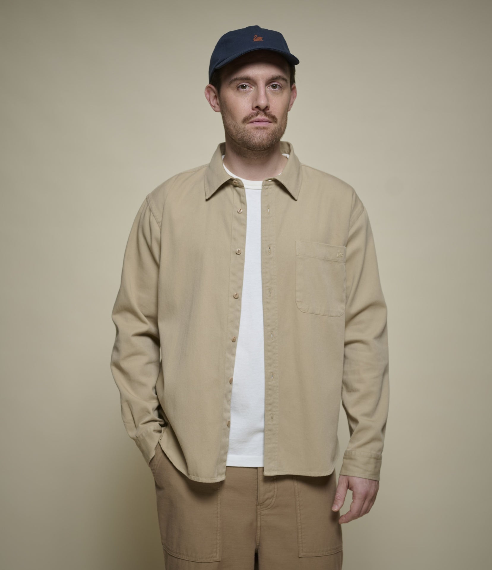 A man wearing a beige long-sleeve button-up overshirt over a white t-shirt, paired with matching beige pants and a navy blue baseball cap, standing against a solid beige background.