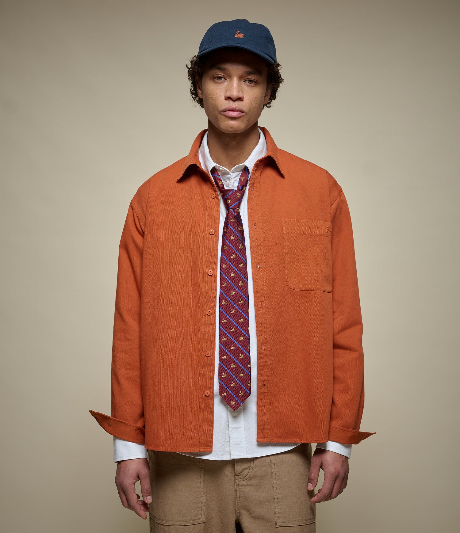 A model wearing an orange button-up overshirt over a white shirt, a patterned purple tie, khaki pants, and a navy blue baseball cap, set against a solid beige background.