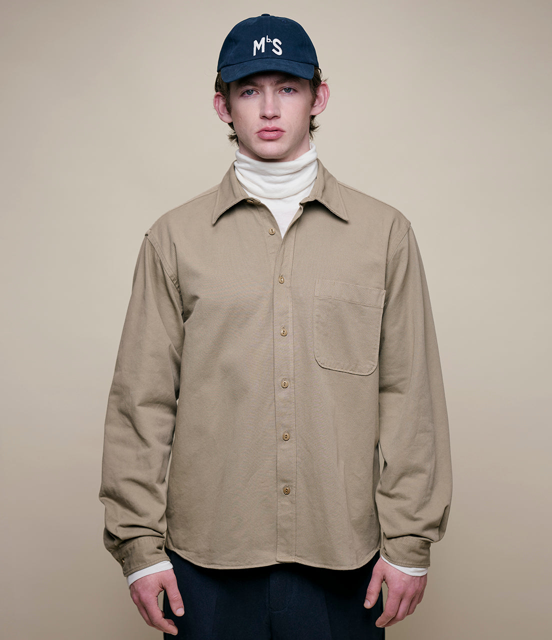 Person wearing a beige twill shirt and navy cap with 'MbS' logo on a beige background