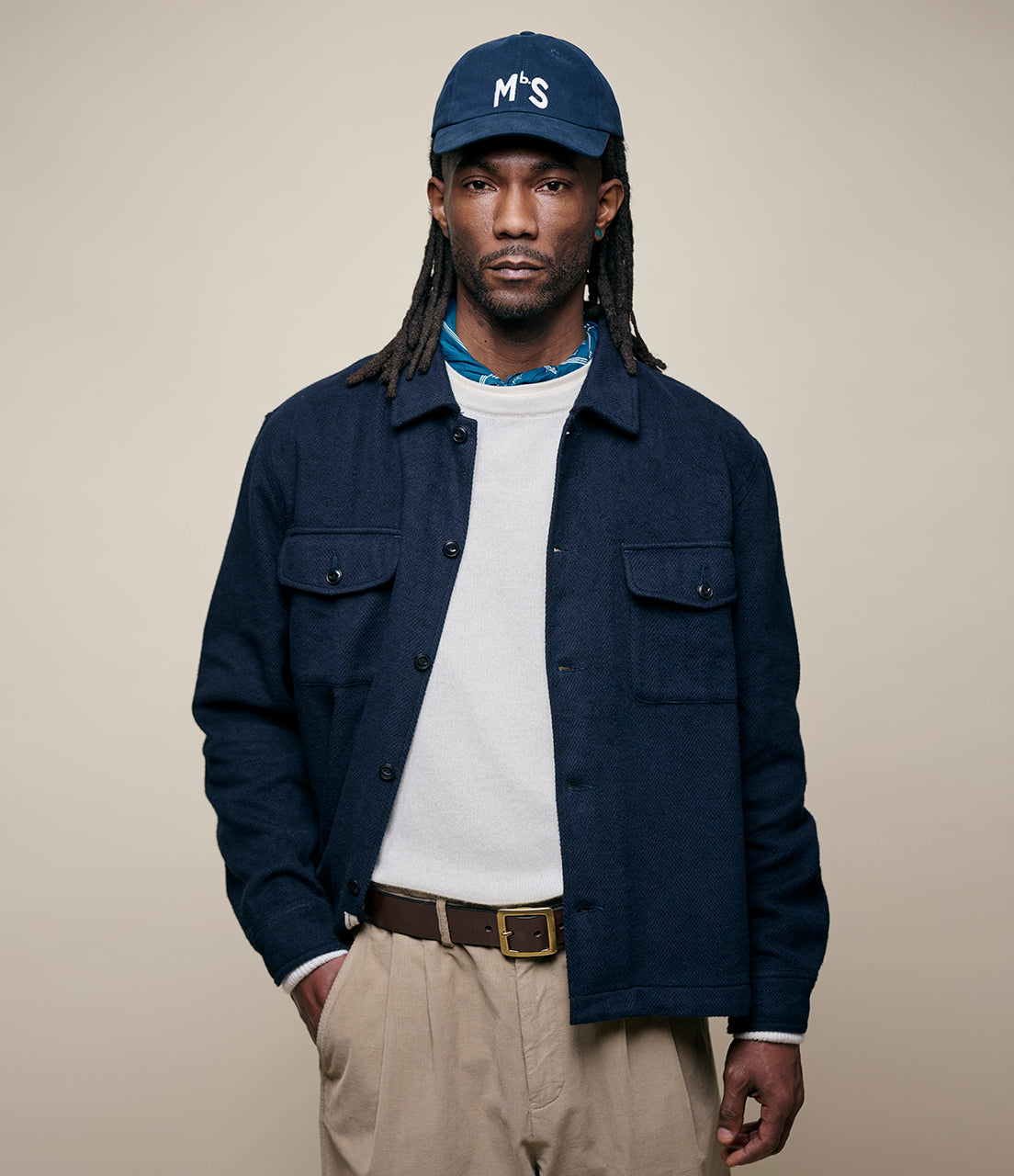 Man wearing a navy flannel shirt, white shirt, beige pants, and navy cap with 'MbS' logo on a beige background