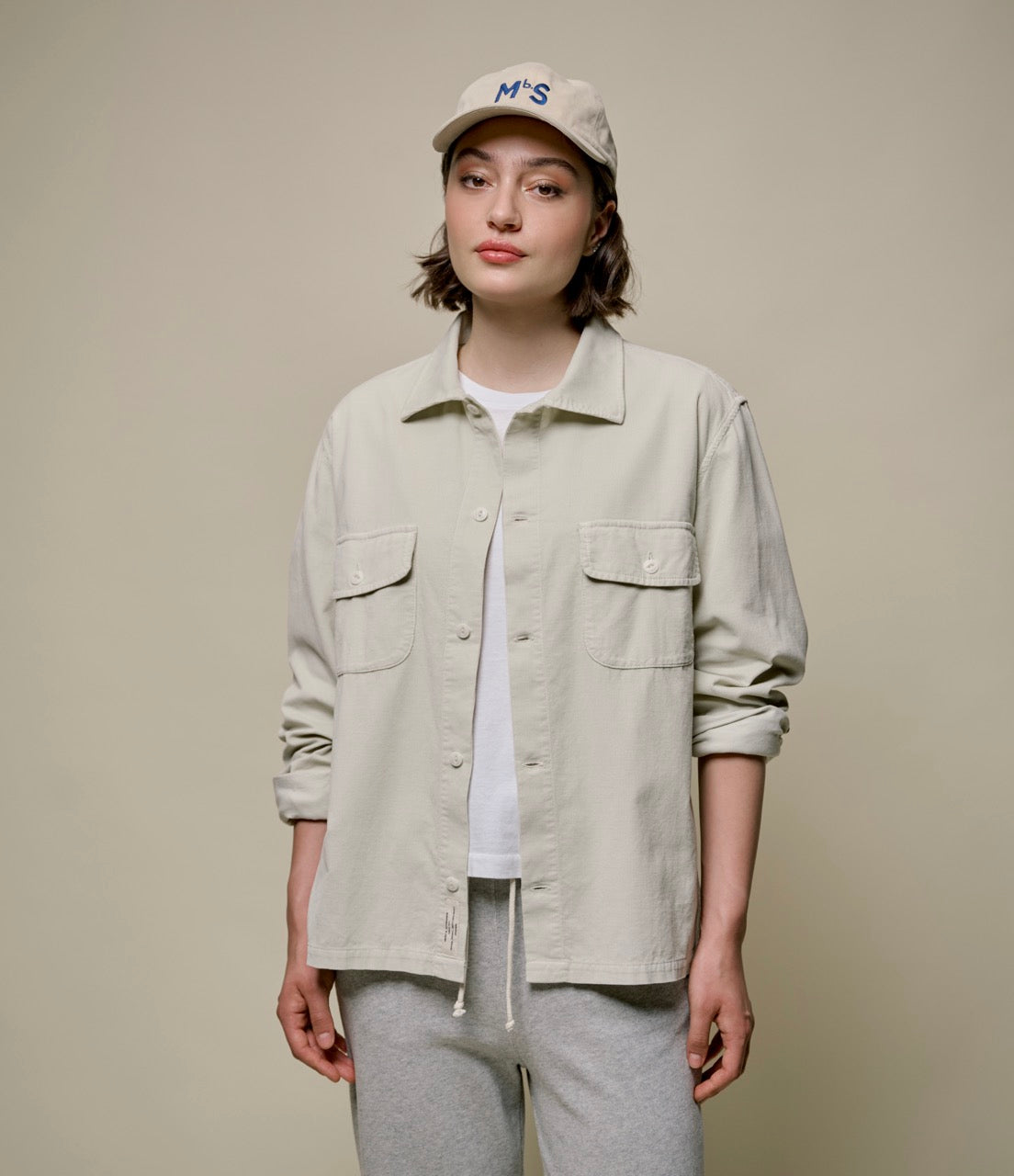 A person wearing a beige long sleeve corduroy shirt with button-up front and collar, paired with light gray pants and a white cap with a logo on the front.