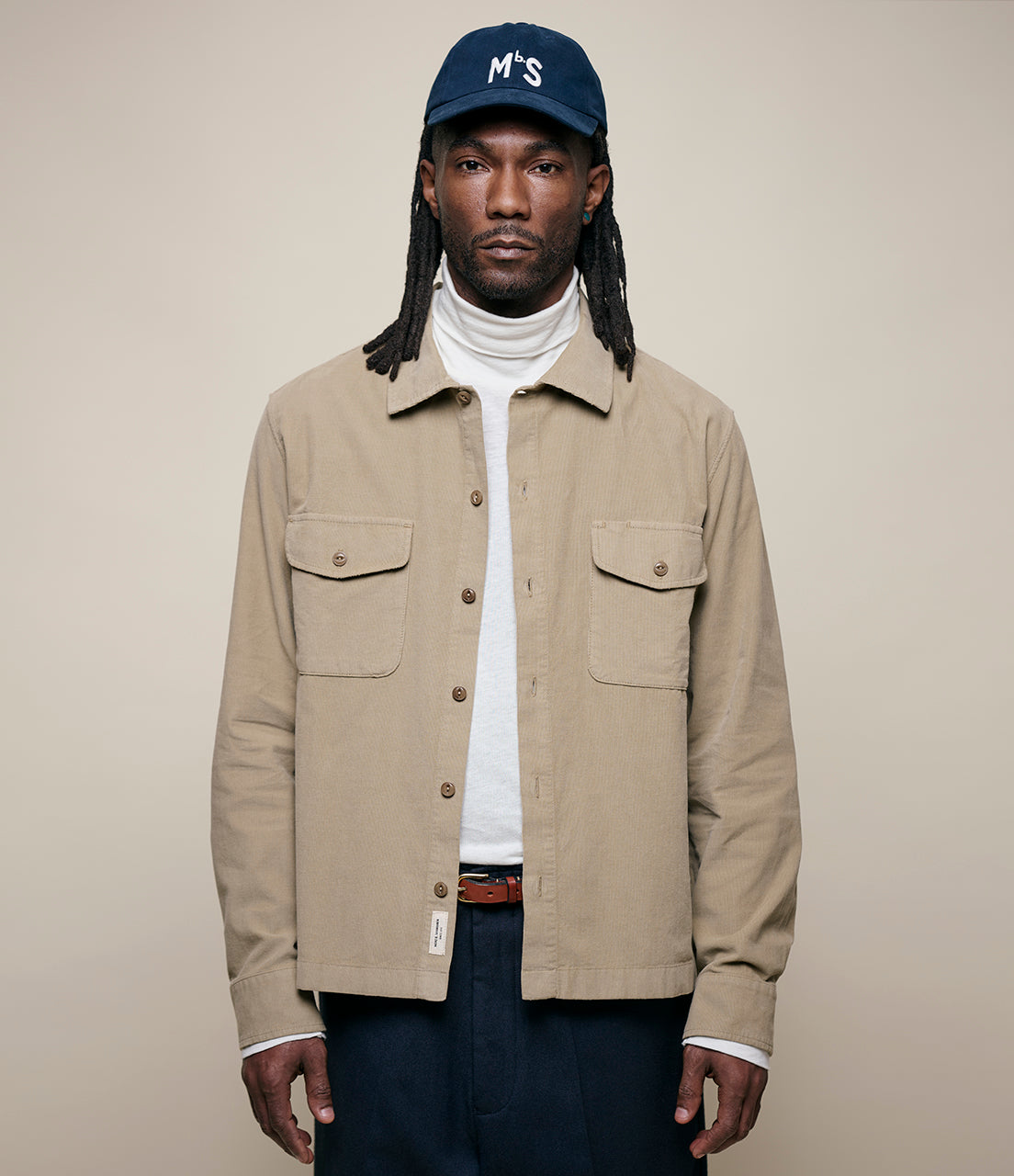 Man wearing a beige corduroy shirt, white turtleneck, and navy cap with 'MbS' logo on a beige background