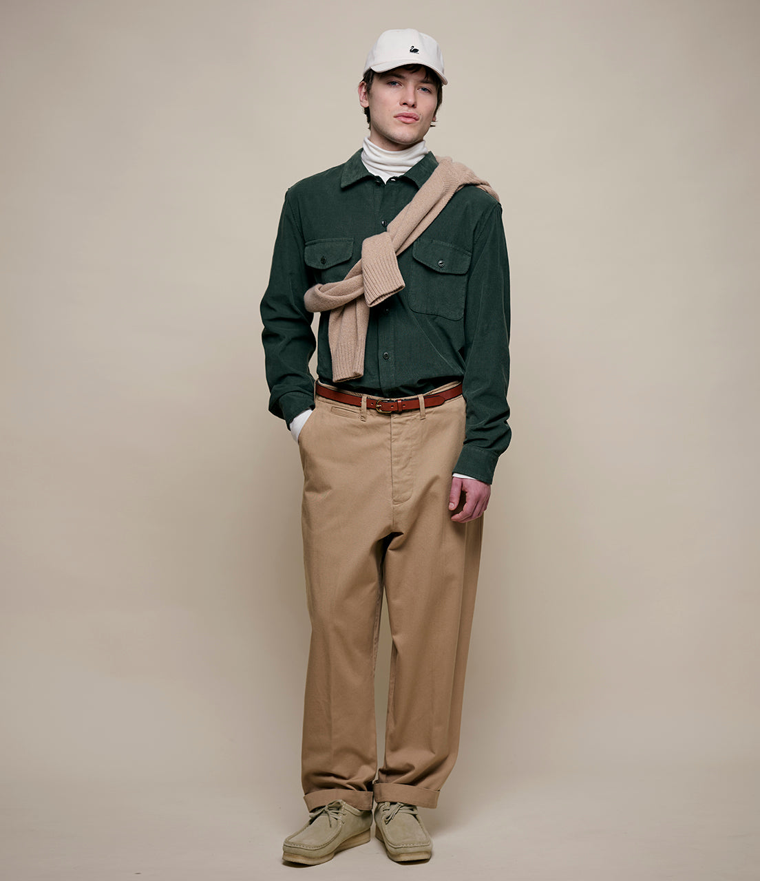 Person wearing a green shirt, beige pants, and a beige scarf against a plain background