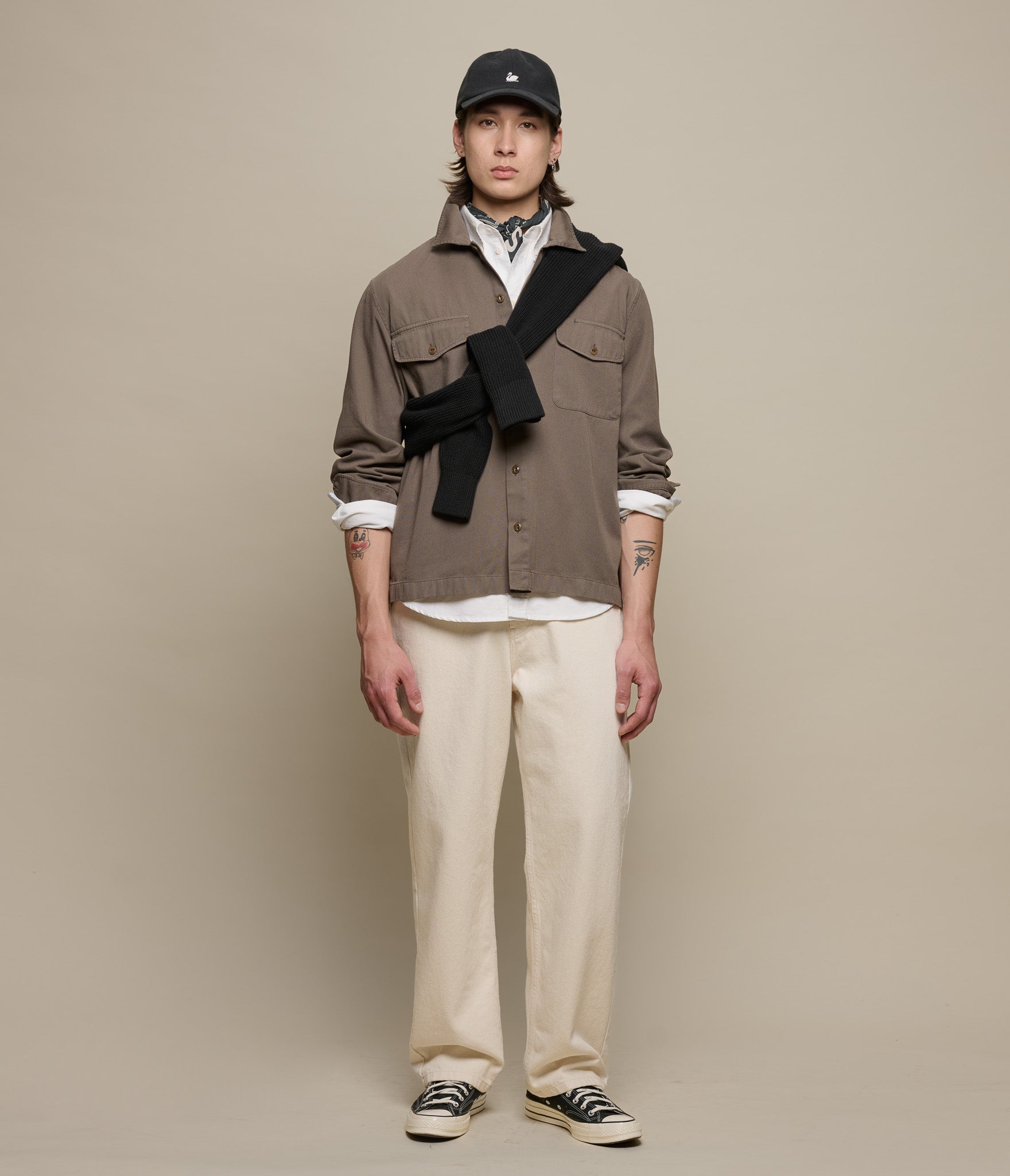 Person wearing a brown jacket, white pants, and black cap against a beige background