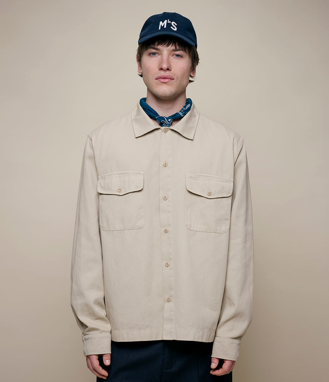 Person wearing a beige jacket and blue cap with 'MbS' logo on a beige background