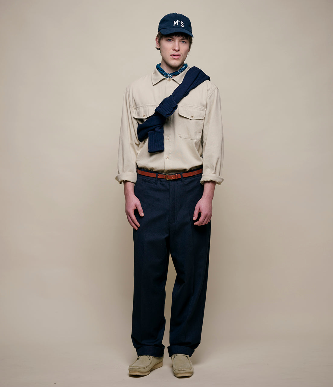 Person wearing a beige jacket, navy pants, and a navy cap on a plain background