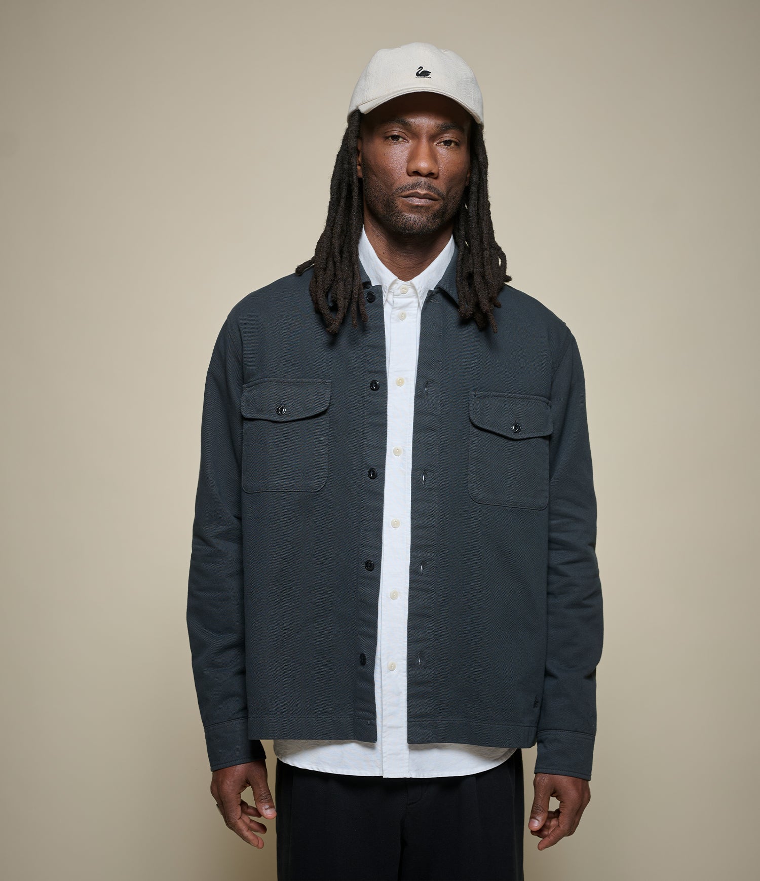 A man wearing a dark charcoal gray utility overshirt over a white button-down shirt, paired with a white baseball cap and black pants, standing against a solid beige background.