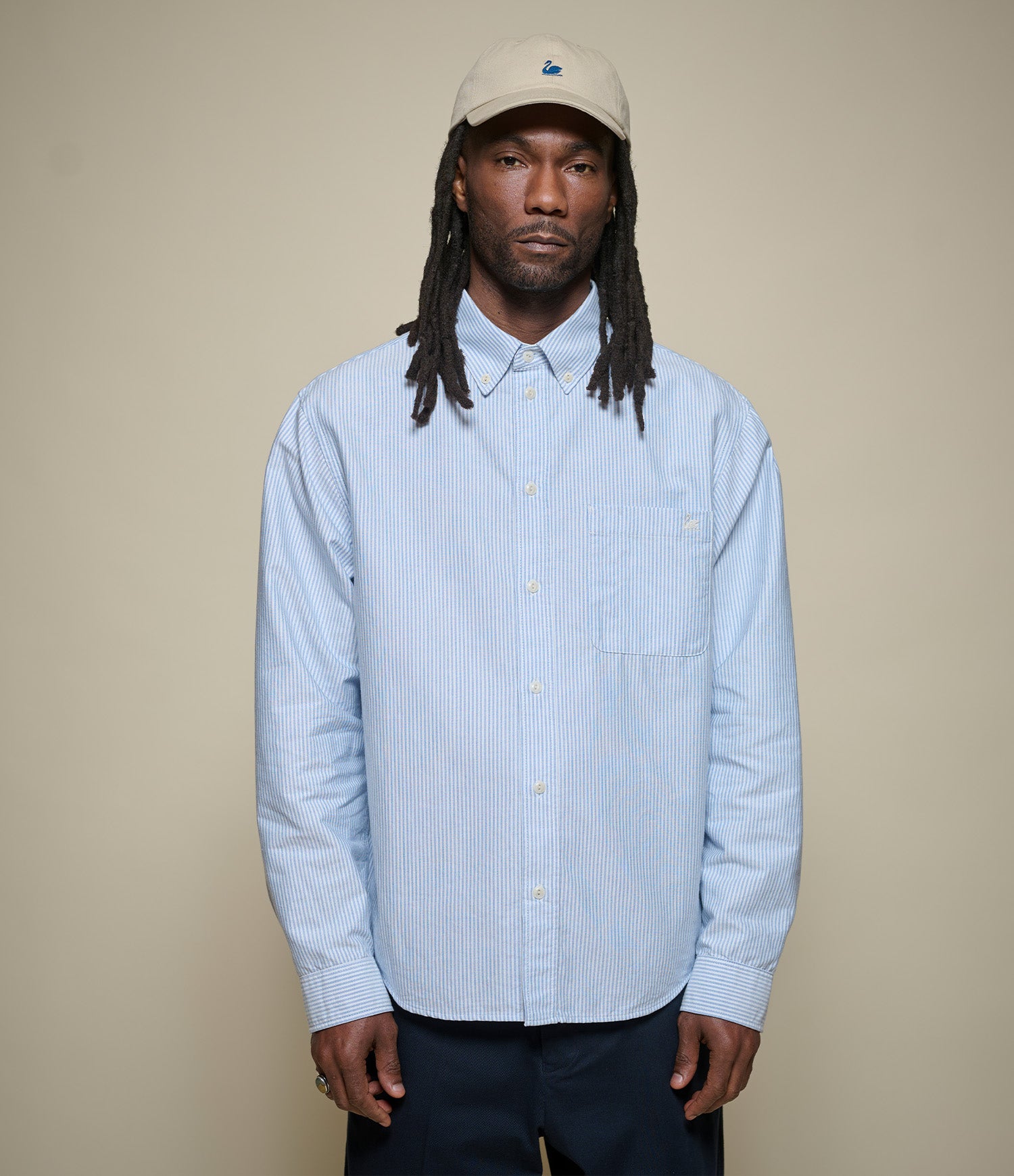 Man wearing a light blue striped button-down shirt and beige cap against a beige background