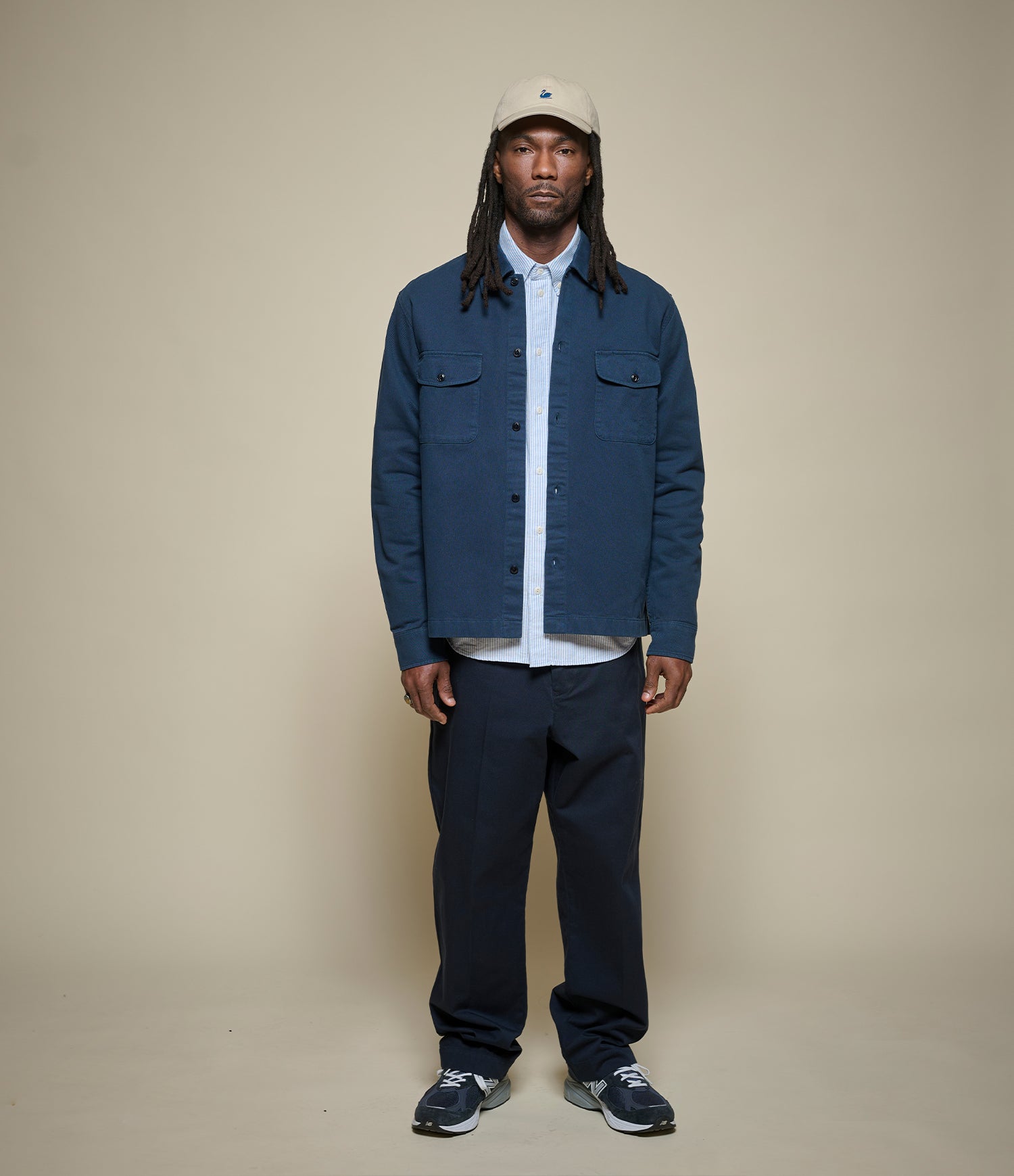 Man wearing a blue jacket, blue striped shirt, dark pants, and a cap on a beige background