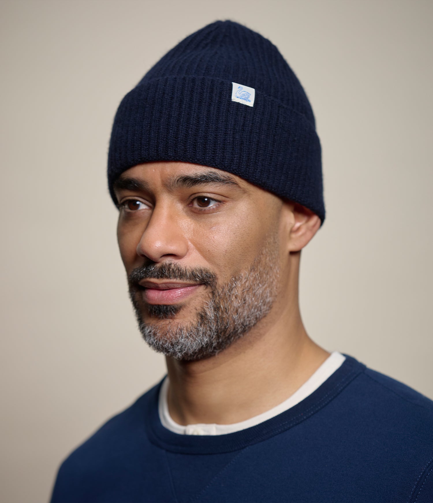 A man wearing a dark navy blue ribbed knit beanie with a small white rectangular logo patch on the side, photographed against a plain beige studio background.