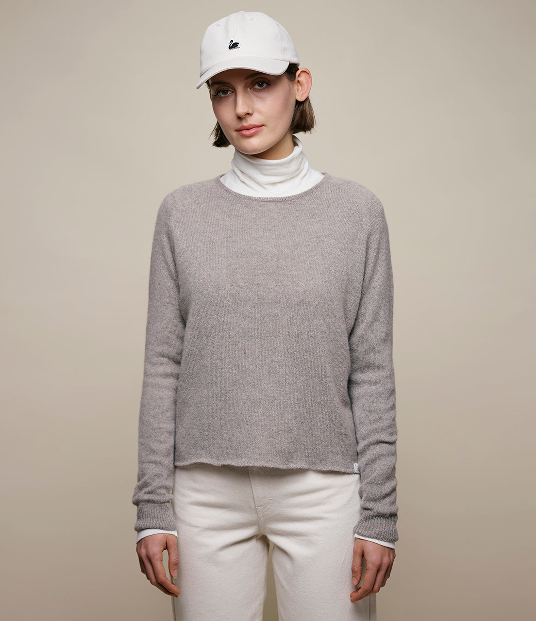 Person wearing a gray sweater, white turtleneck, and white pants with a beige background