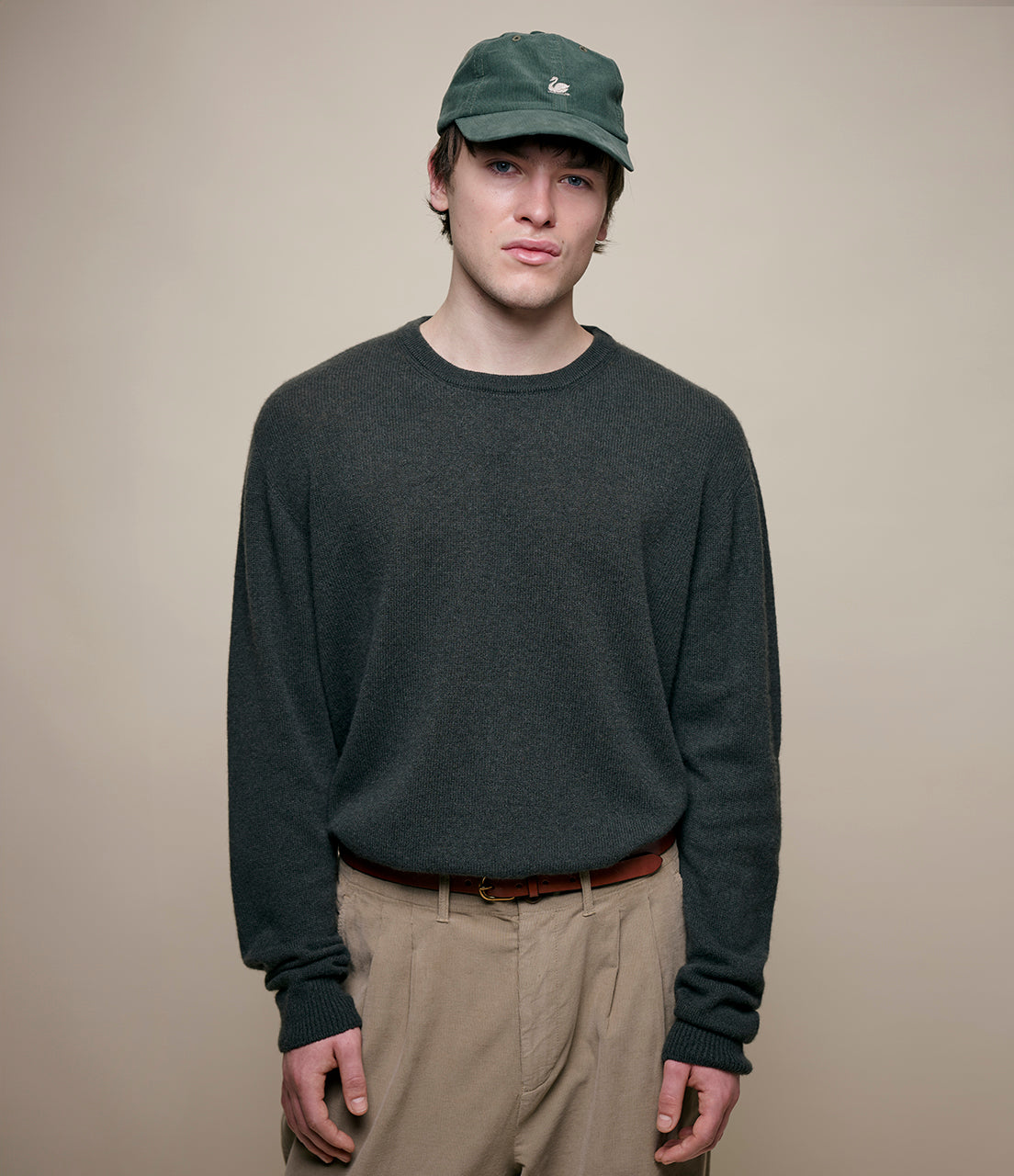 Person wearing a dark green sweater, beige pants, and a green cap against a plain background