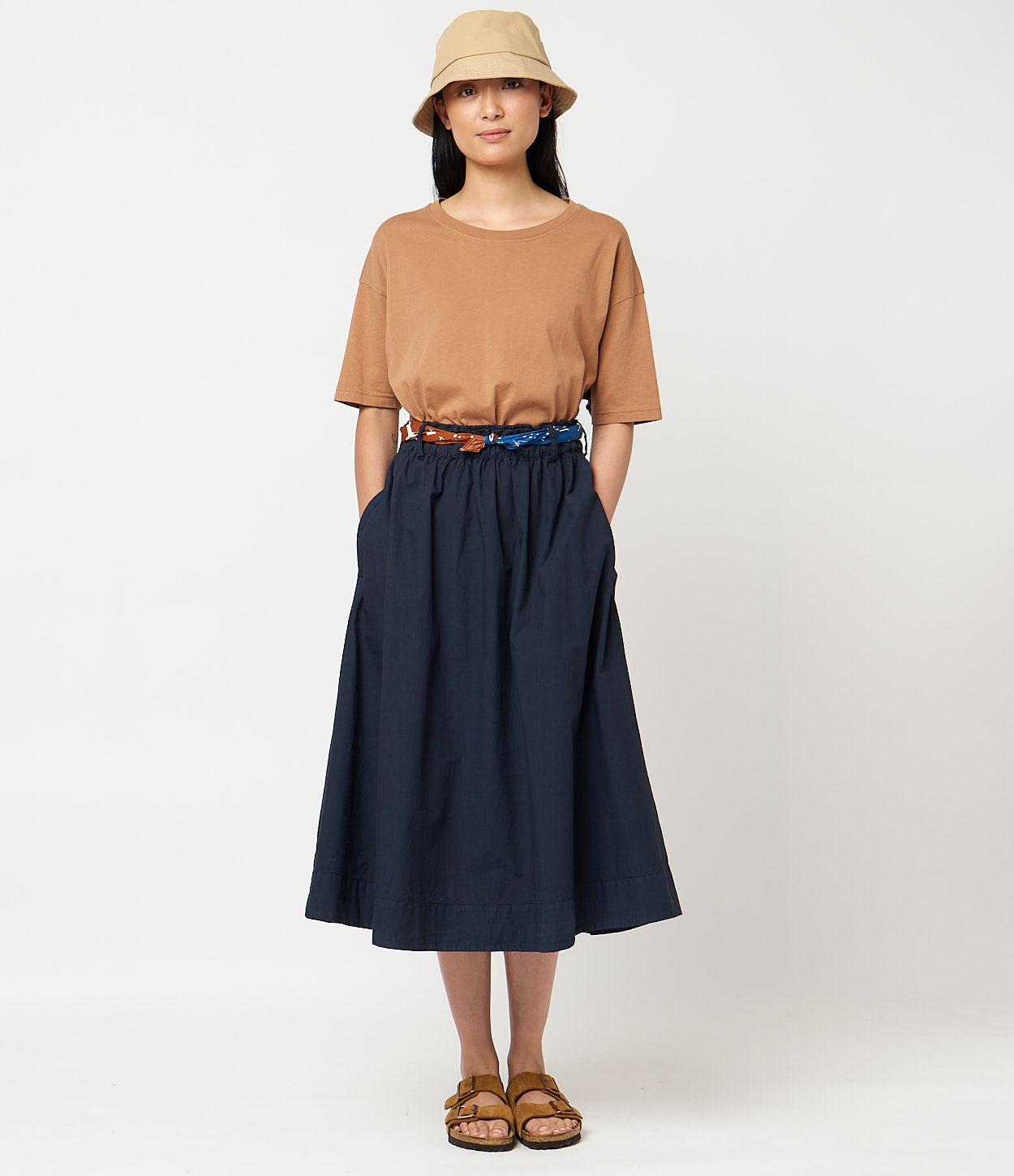 A model wearing a beige top and a navy flared cotton poplin skirt with a wide waistband, paired with brown sandals and a wide-brimmed hat.