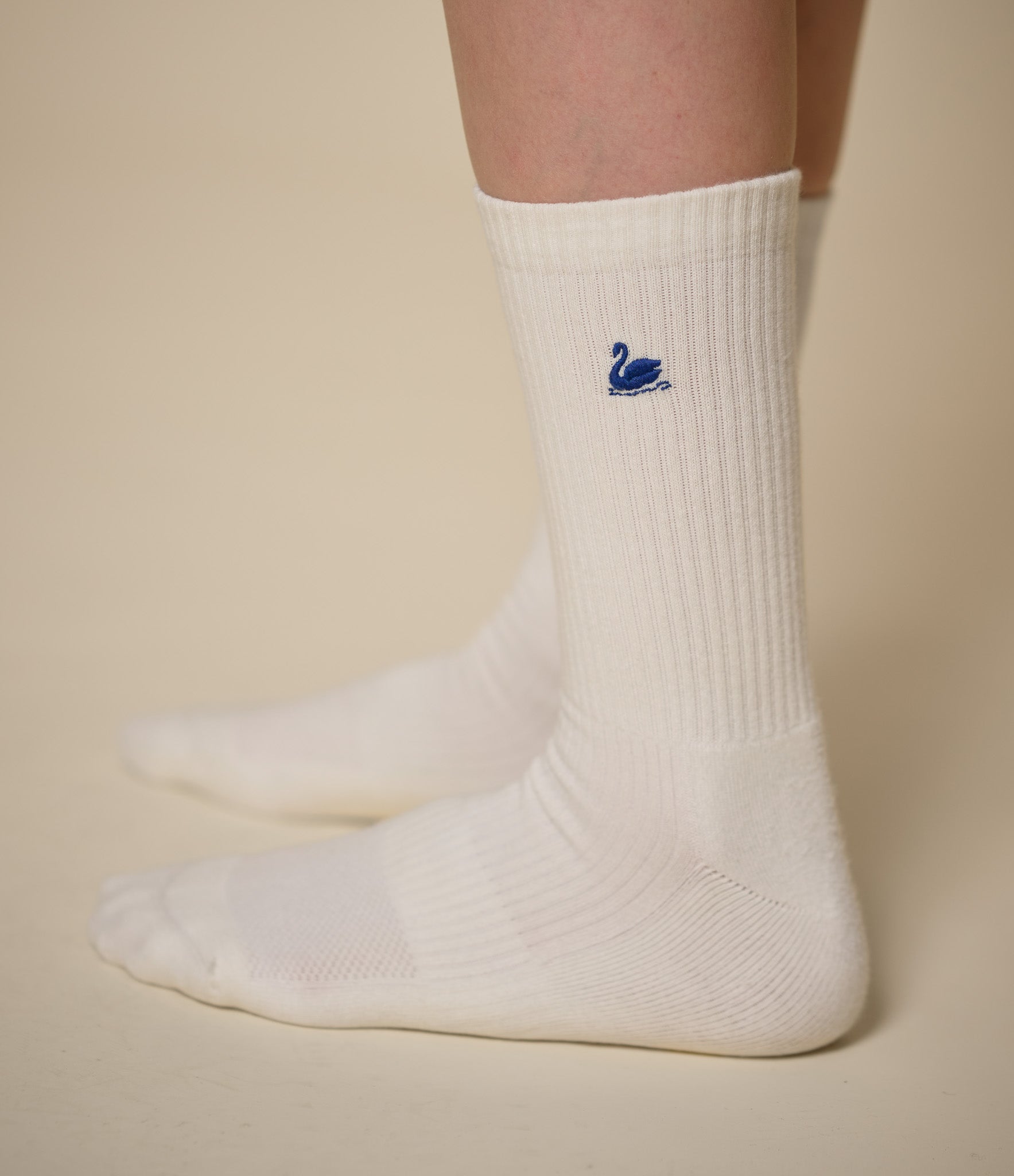 A pair of white ribbed crew socks worn on feet, featuring a small blue embroidered swan logo on the side, set against a solid beige background.