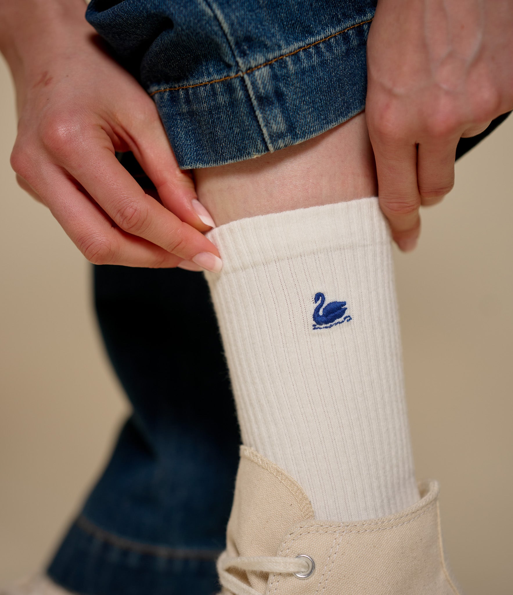 A close-up of a person's leg and hands adjusting a ribbed white crew sock with a small navy blue swan logo, paired with a beige ankle boot and blue denim jeans.
