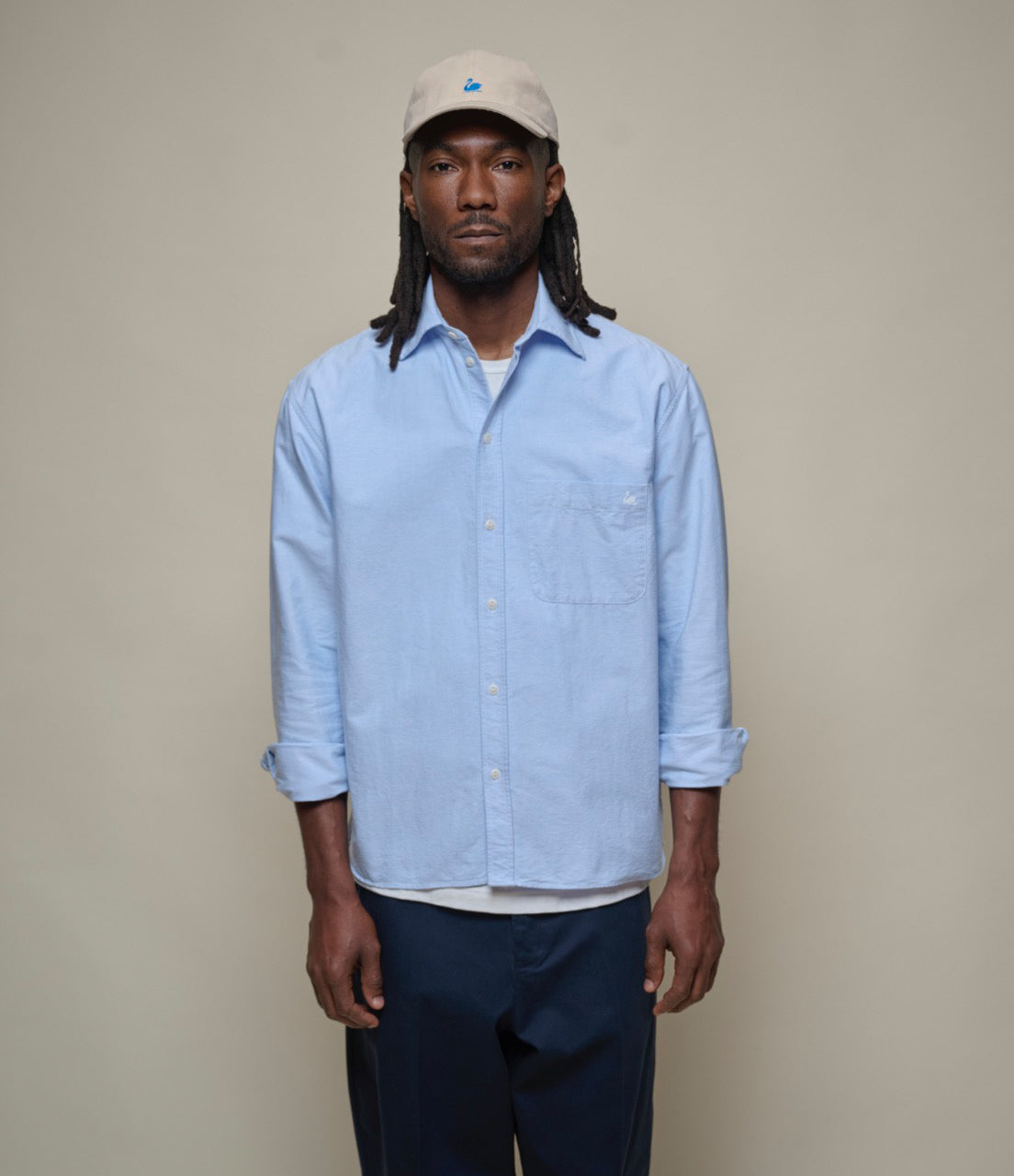 Man wearing a light blue  oxford shirt and beige cap against a plain background