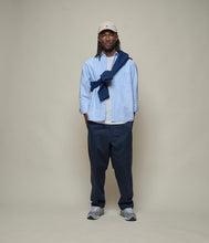 Man wearing blue oxford shirt over a white T-shirt, with navy blue chinos and beige cap