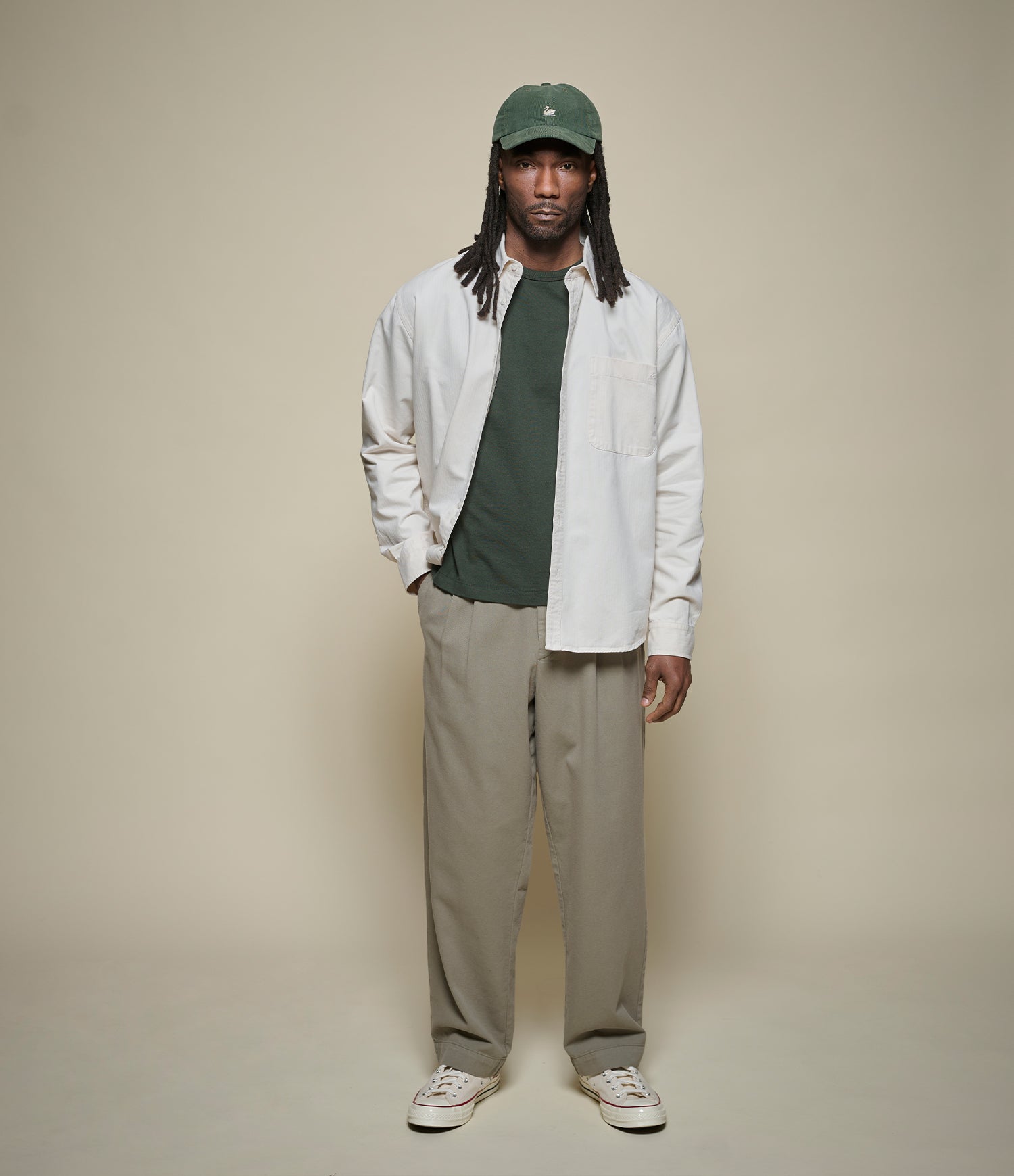 Man wearing a white shirt over a cropped green T-shirt, green cap, and green pleated pants against a beige background