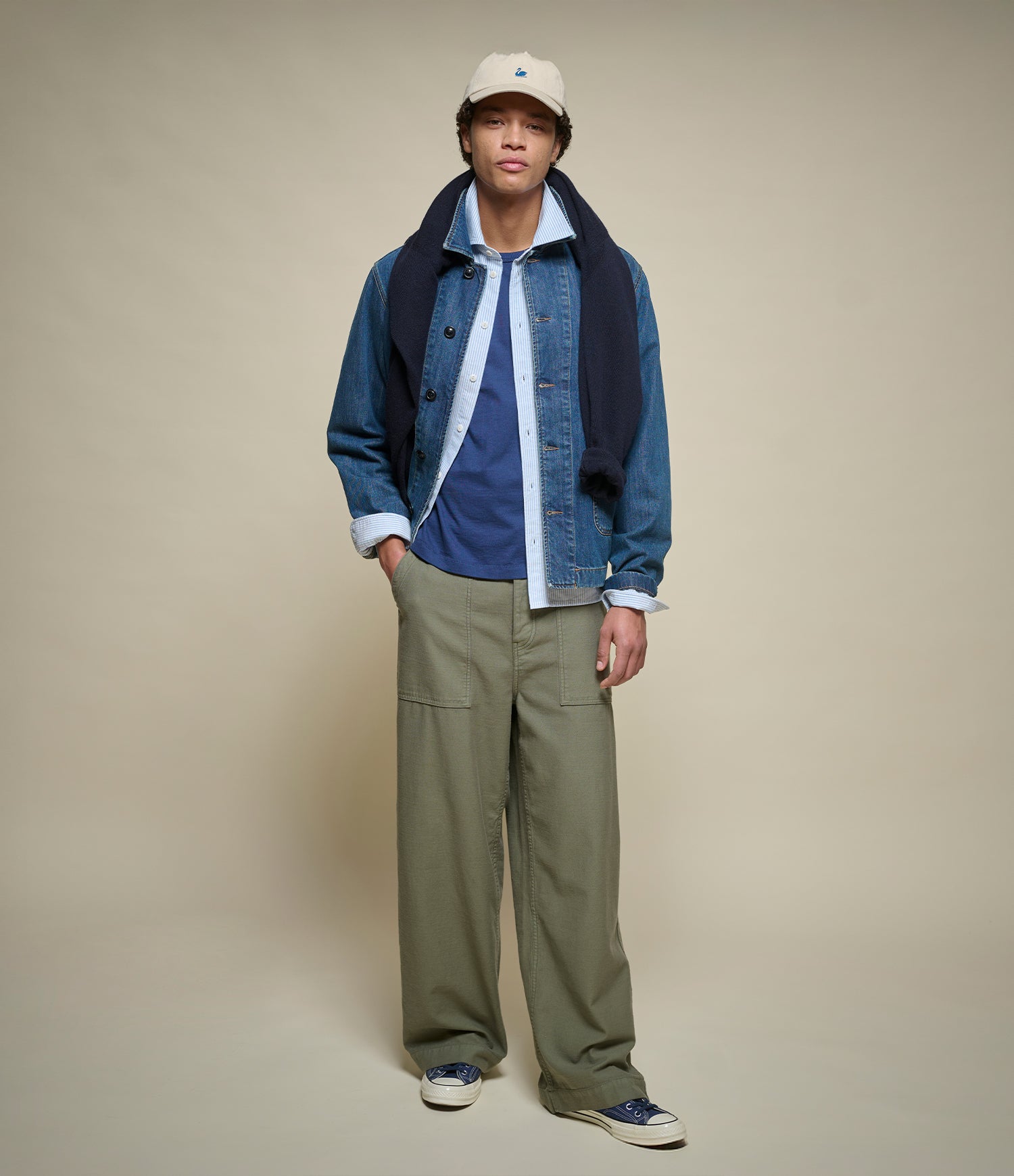 Person wearing a blue denim jacket, blue oxford shirt, blue t-shirt, green pants, and a beige cap on a beige background