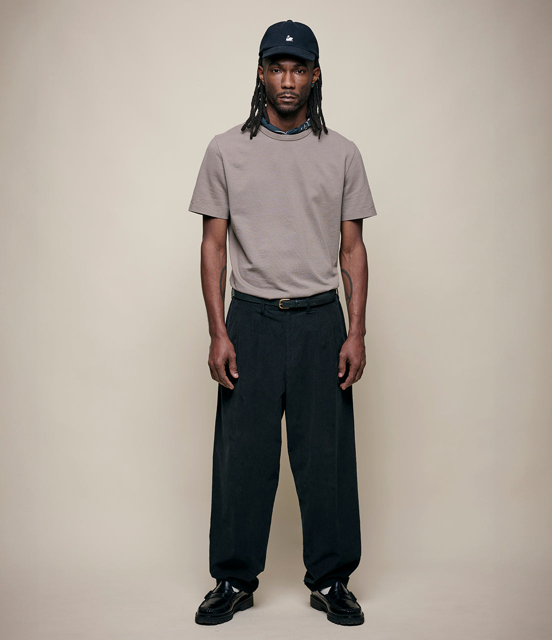 Man wearing a gray t-shirt, black pants, and a black cap against a beige background
