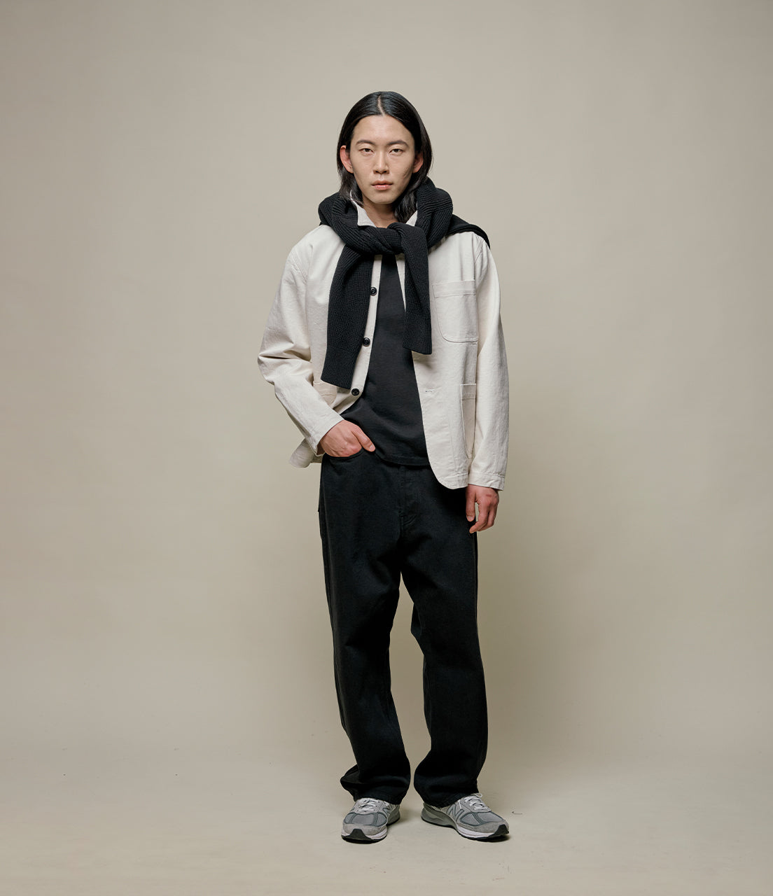 Person wearing a white jacket, black pants, and a dark scarf against a beige background