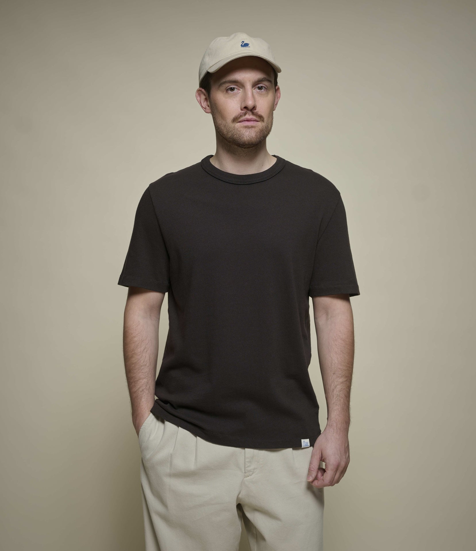 Man wearing a dark brown crew neck t-shirt and beige cap against a beige background