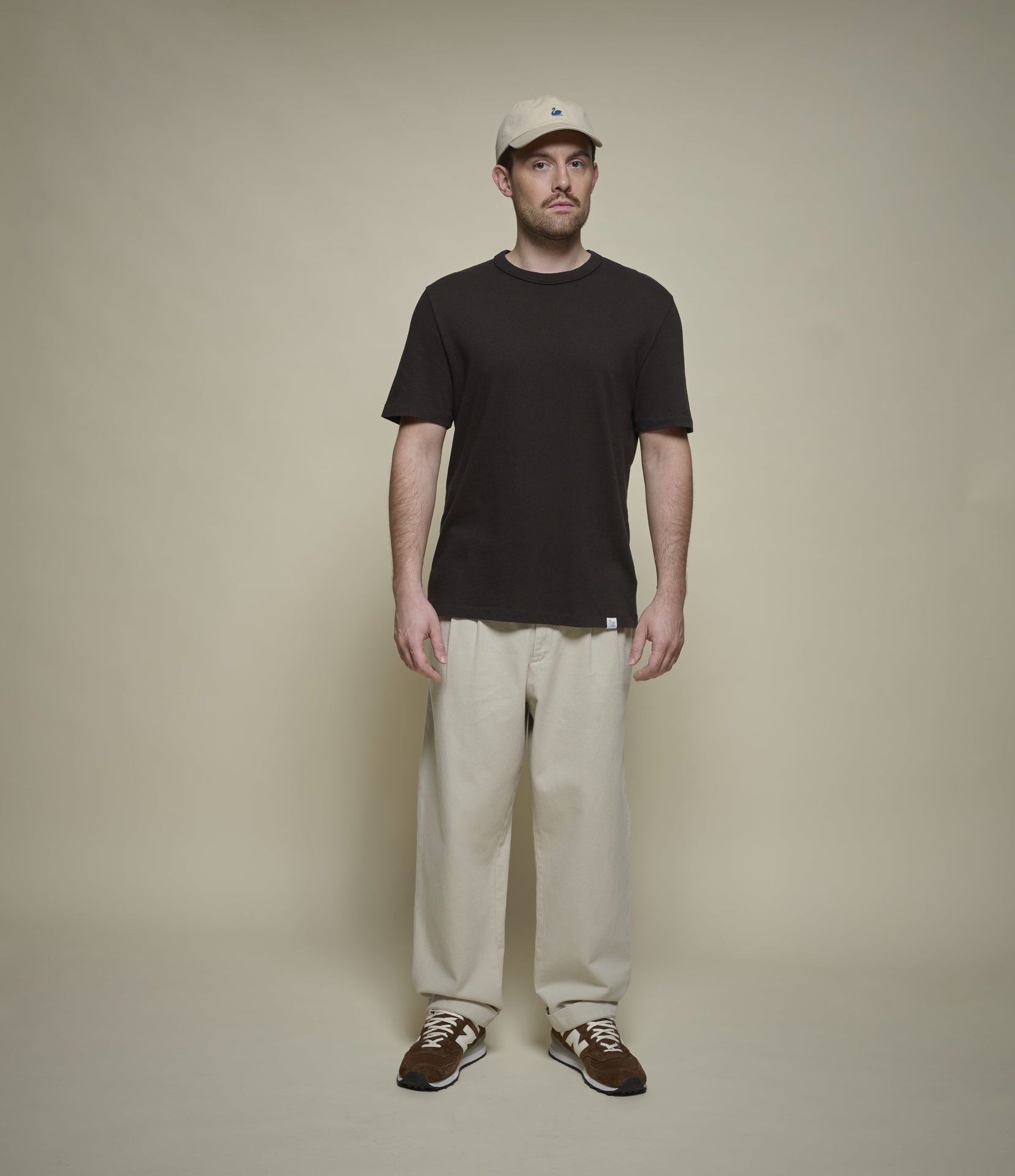 Man wearing a dark brown t-shirt, beige pants, and a cap against a plain background