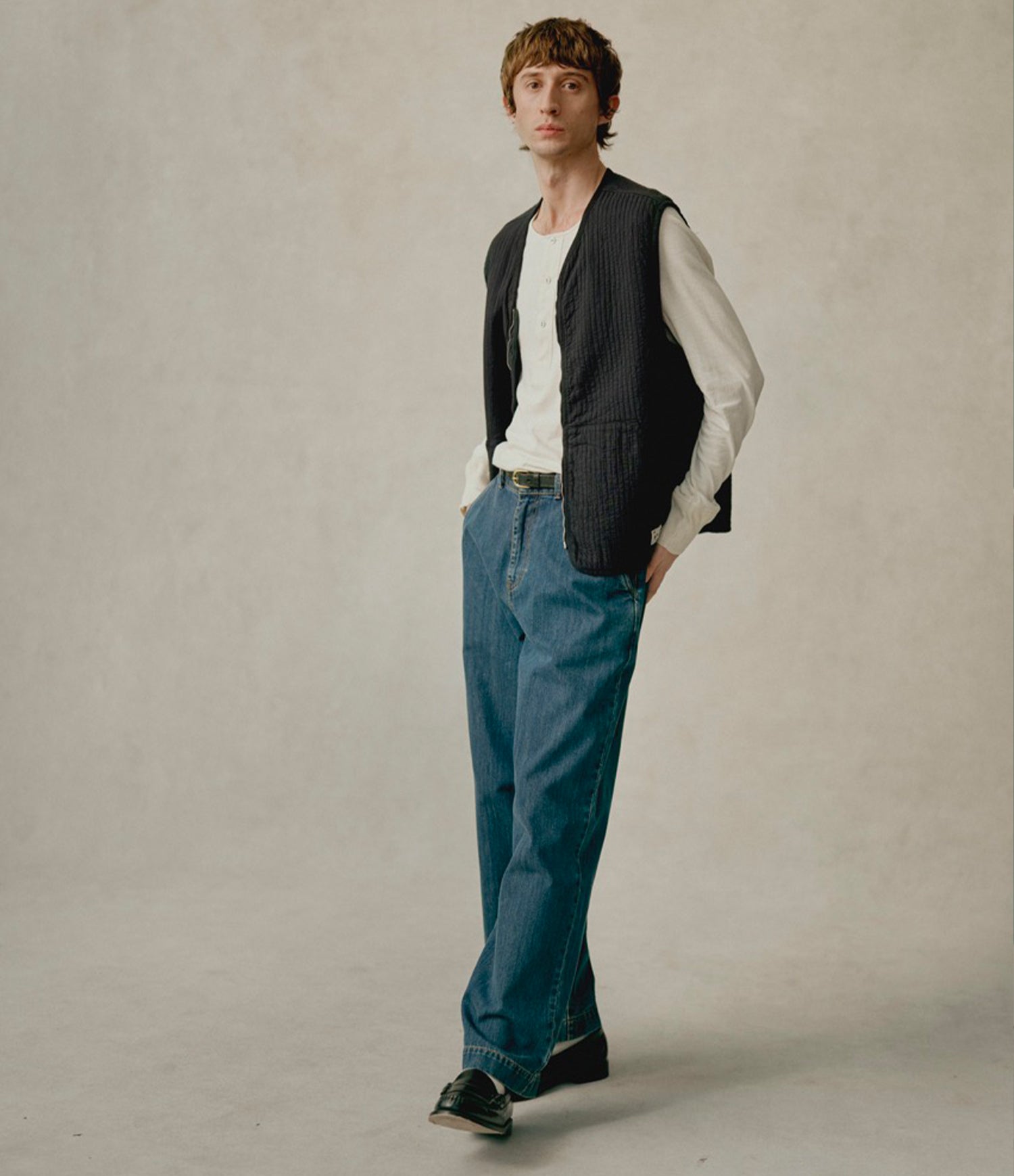 A man stands in a studio wearing a black quilted vest over a white long-sleeve shirt, blue jeans, and black sneakers against a plain light gray background.